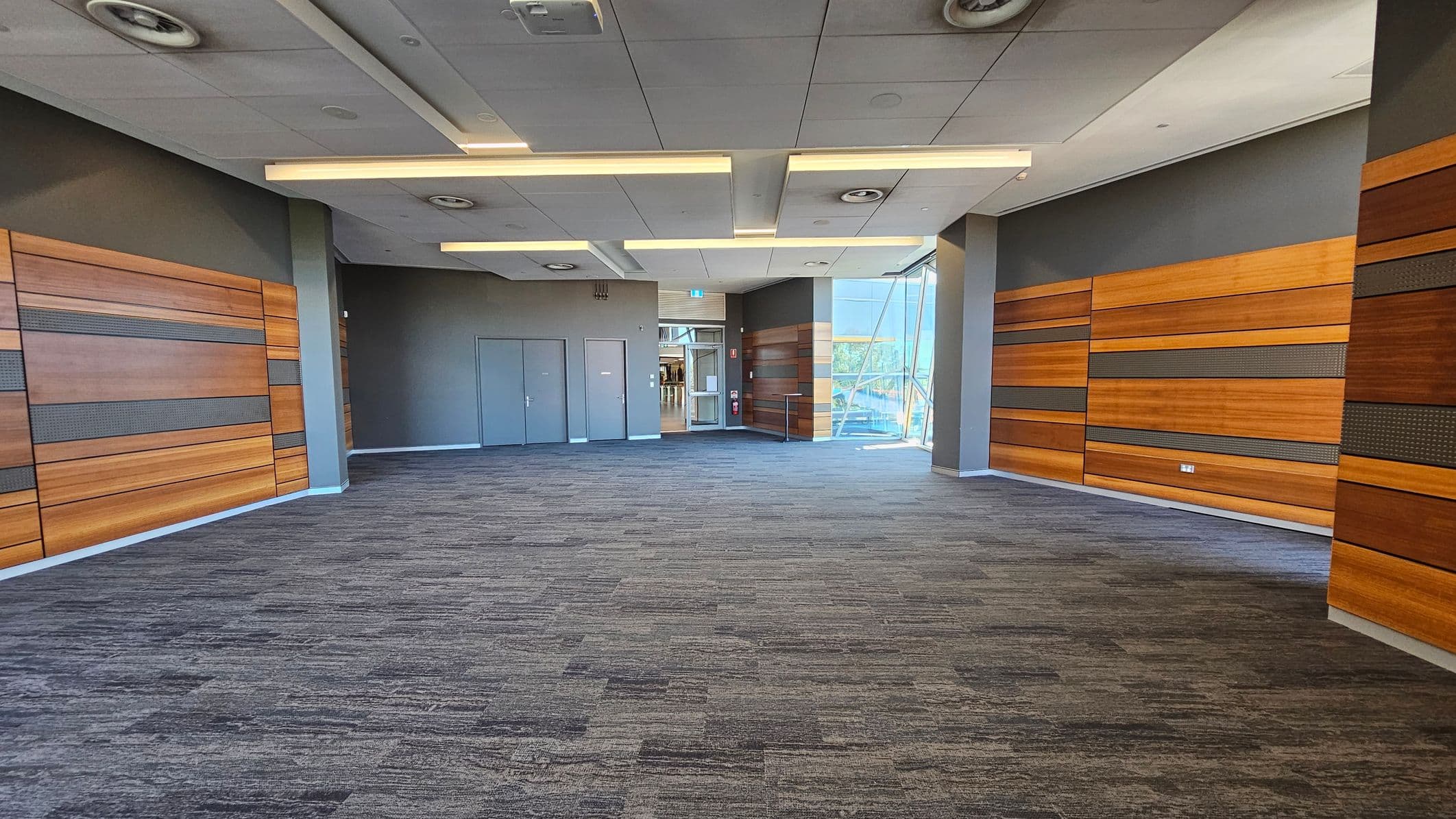 A spacious room with a grey carpeted floor and a ceiling featuring recessed lighting and circular vents. The walls are adorned with horizontal wooden panels in varying shades. On the left, there are two grey doors. At the far end, a glass entrance with a view of a staircase and outdoor greenery. The room is well-lit with natural light streaming in from the glass area.