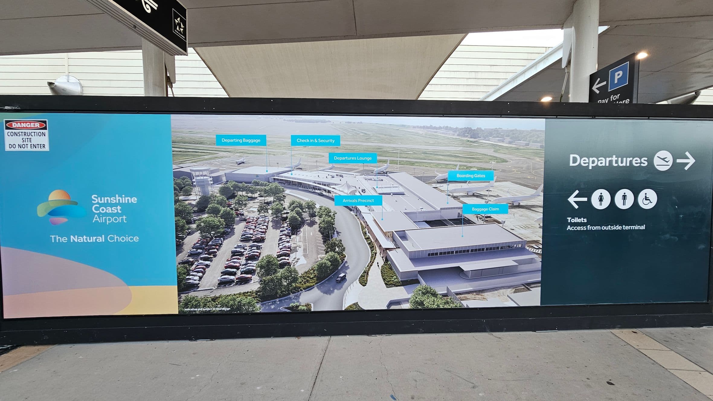 A large sign at Sunshine Coast Airport. On the left, a blue section with the airport's logo and the slogan "The Natural Choice." Above it, a "Danger Construction Site Do Not Enter" warning. In the middle, an aerial view of the airport with labels for various areas like "Departing Baggage" and "Arrivals Precinct." On the right, a dark section with "Departures" and icons for toilets, including accessibility symbols. The floor is grey concrete, and the ceiling has a modern design with beams.