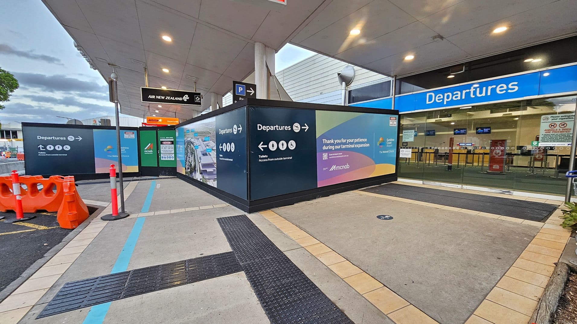 A covered walkway leads to the departures area of an airport. On the left, there are orange traffic barriers and signs directing passengers to departures and toilets. The right side features a glass entrance with a blue "Departures" sign above. The floor is a mix of concrete and tiled sections, with a blue line marking a path. The ceiling is white with recessed lighting. A construction notice thanks passengers for their patience during terminal expansion.
