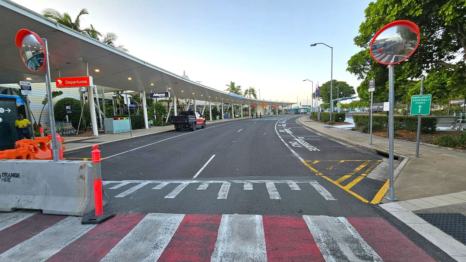 A road with a pedestrian crossing in the foreground, marked with red and white stripes. On the left, there are orange traffic cones and a concrete barrier. A covered walkway with signs for "Departures" and "Alliance" runs along the left side, with a black vehicle parked nearby. On the right, a convex mirror is mounted on a pole, and a green sign indicates an "Emergency Assembly Point." Trees and bushes line the pavement, and the road curves to the right in the background.