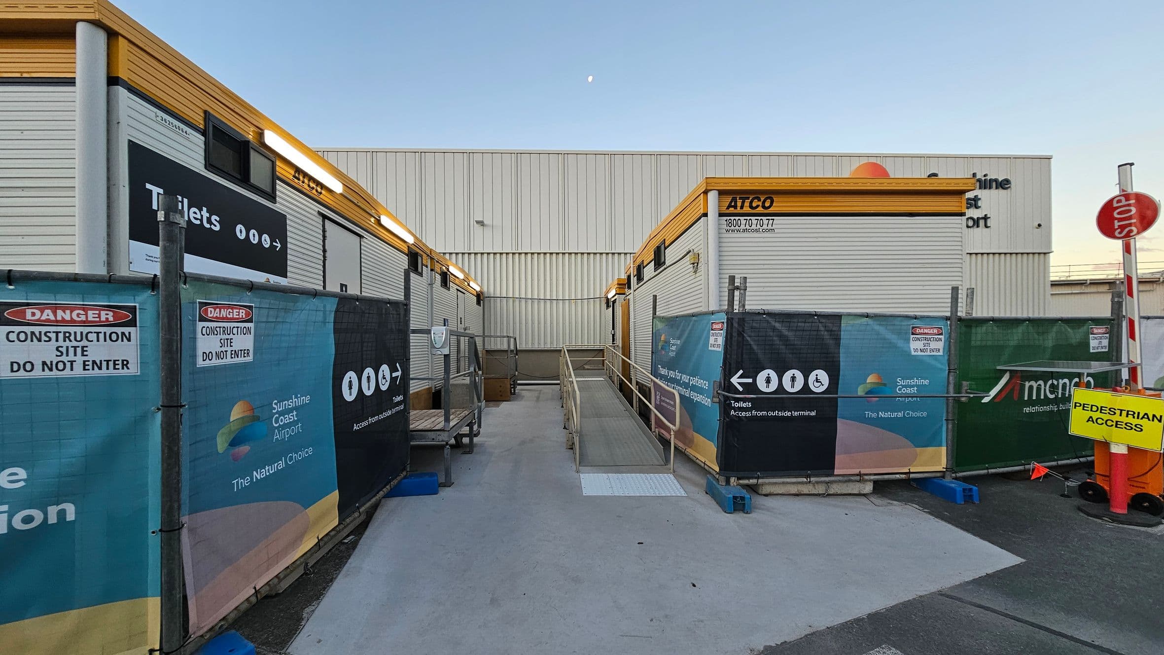 Construction site with portable buildings on either side, marked "ATCO" with yellow trim. Signs indicate toilets and pedestrian access. Fencing displays "Danger Construction Site" and "Sunshine Coast Airport" branding. A ramp leads to the buildings. A "Stop" sign and a yellow "Pedestrian Access" sign are visible. The ground is concrete, and the background shows a large white building with partial signage.