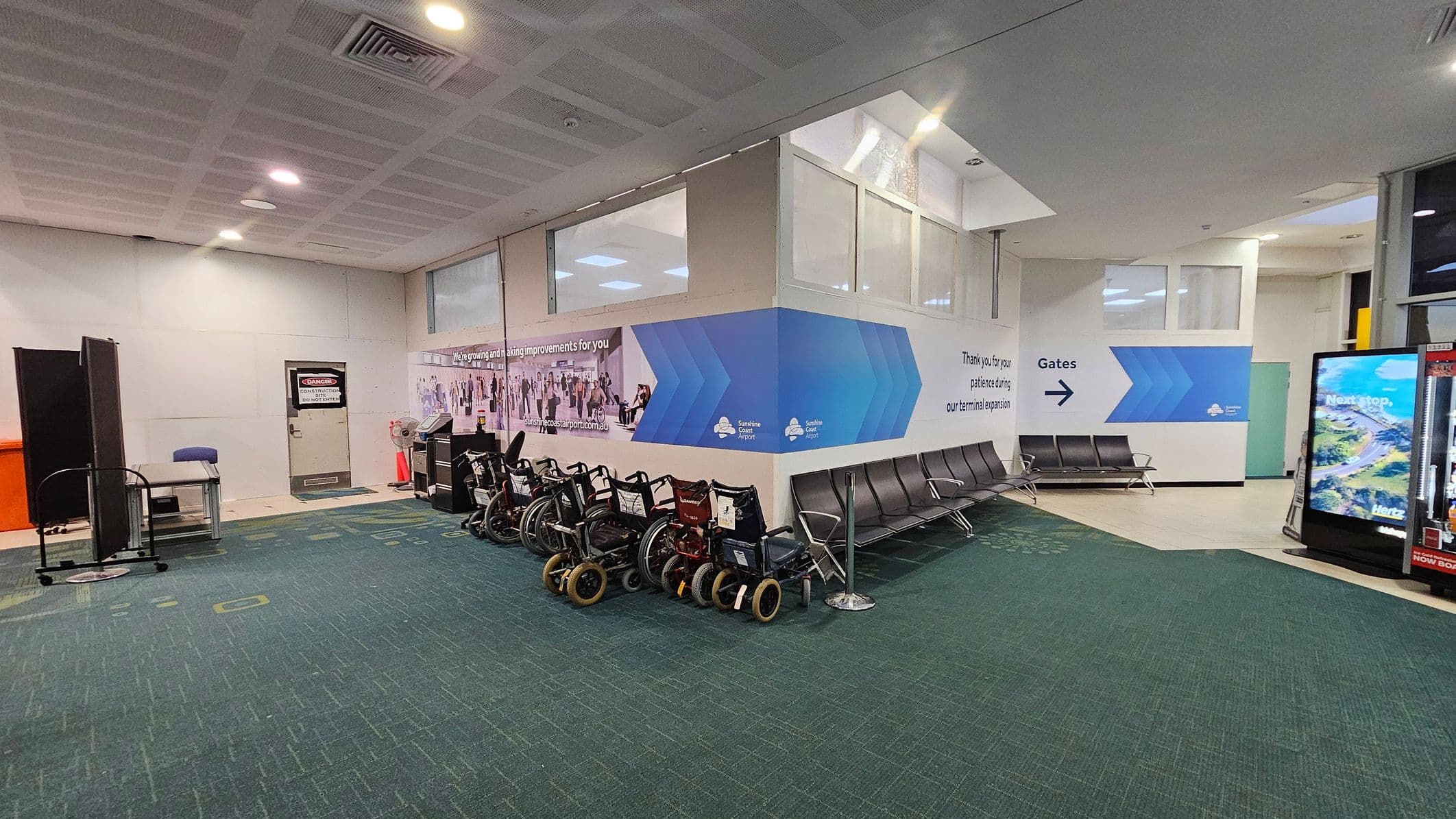 A waiting area in an airport with a green carpeted floor. On the left, there is a door with a sign and a small desk with a chair. Several wheelchairs are lined up against the wall. In the centre, a wall displays signage about terminal expansion. On the right, there are rows of black seating and a digital display screen. The ceiling is white with recessed lighting and air vents.