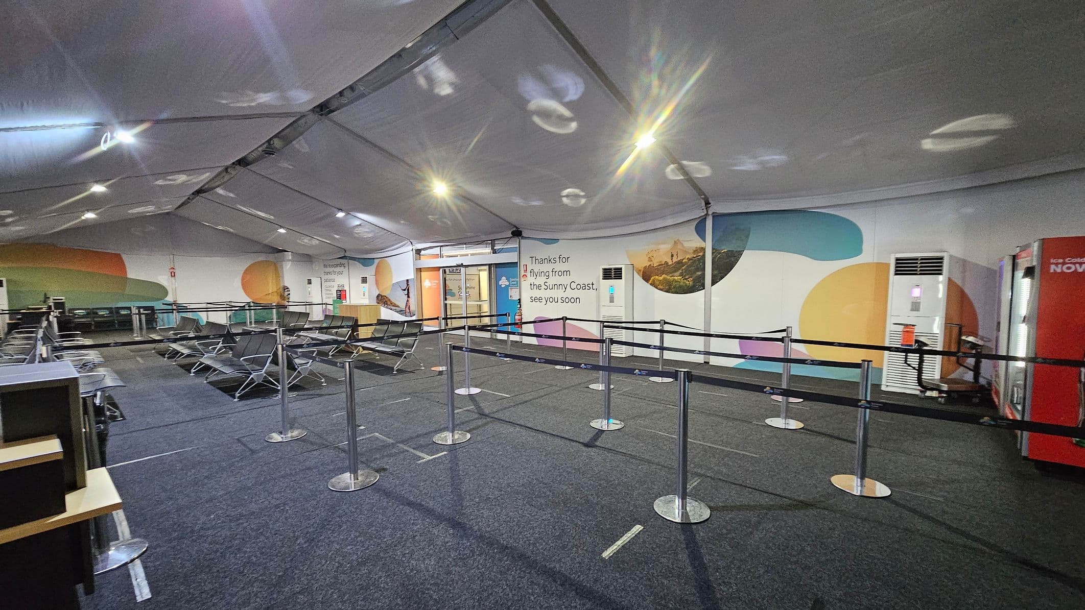 A spacious indoor area with a high, white tent-like ceiling. The floor is carpeted in dark grey. On the left, there are rows of metal and black mesh seating. In the centre, there are queue barriers with metal posts and black straps. The right side features a colourful wall with abstract shapes and a message thanking travellers. An air conditioning unit and a red vending machine are also on the right. The lighting is bright, with reflections visible on the ceiling.