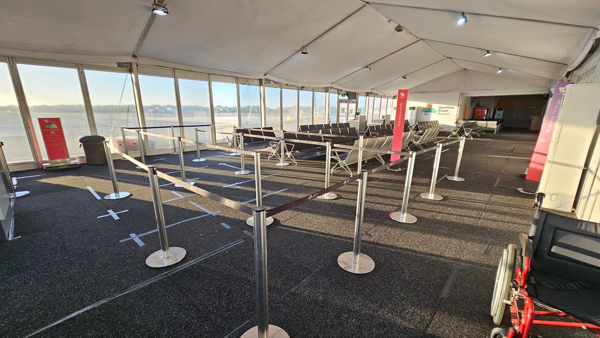 Inside an airport terminal with a carpeted floor and a white tent-like ceiling. On the left, large windows offer a view of the outside, with a red sign and a bin nearby. In the centre, there are rows of metal and black seating, with queue barriers forming a pathway. On the right, a red wheelchair is partially visible. The background features a sign for Sunshine Coast Airport and more seating. Overhead lights are evenly spaced along the ceiling.