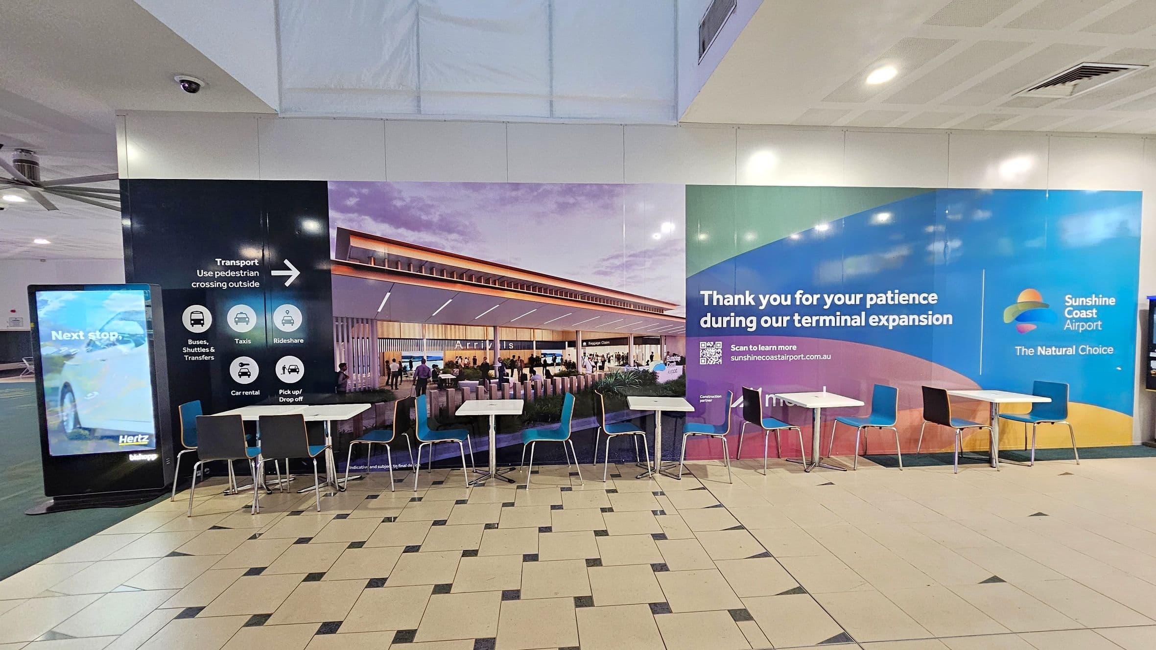 A tiled floor with a pattern of light and dark squares. In the foreground, there are several tables with blue and black chairs. The background features a large mural depicting an airport terminal and signage for Sunshine Coast Airport, thanking visitors for their patience during terminal expansion. On the left, there's a digital display with transport information and a Hertz advertisement. The ceiling is white with recessed lighting and a visible air vent.