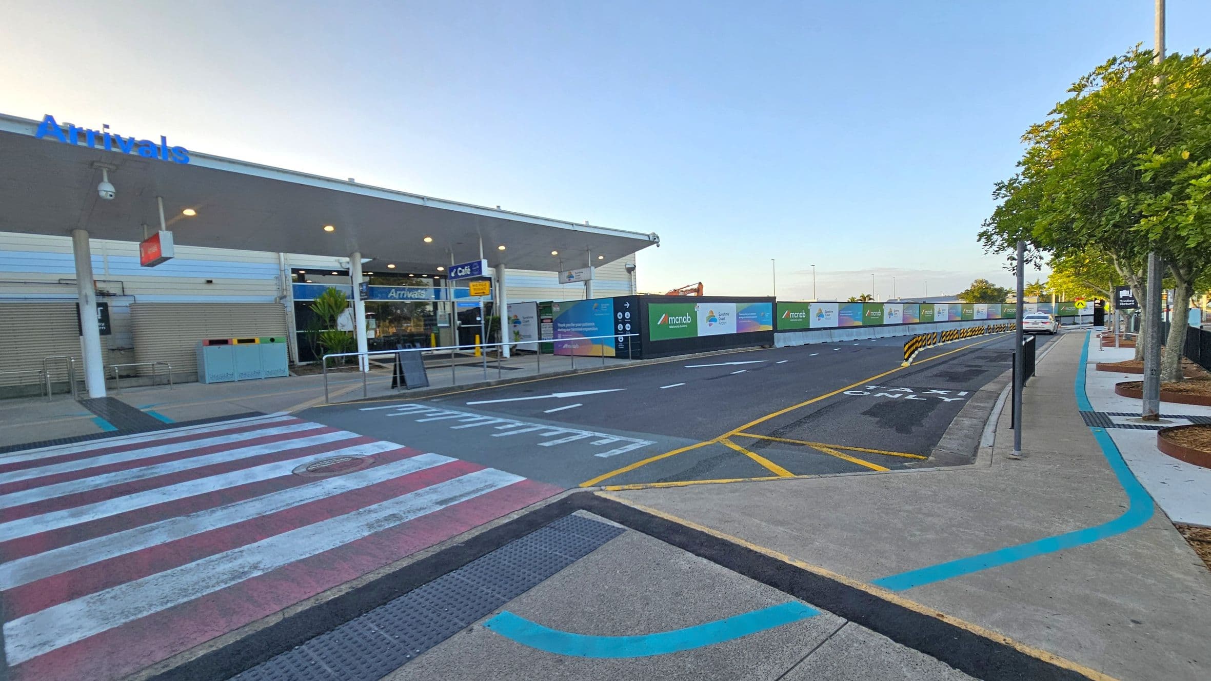 A modern airport arrivals area with a covered walkway on the left, featuring a sign that reads "Arrivals" in blue. The entrance is glass-fronted with visible signage for a café. A pedestrian crossing with red and white stripes is in the foreground. On the right, a road with marked lanes runs parallel to a pavement lined with trees. Construction barriers with advertisements are visible in the background, along with a parked car further down the road.