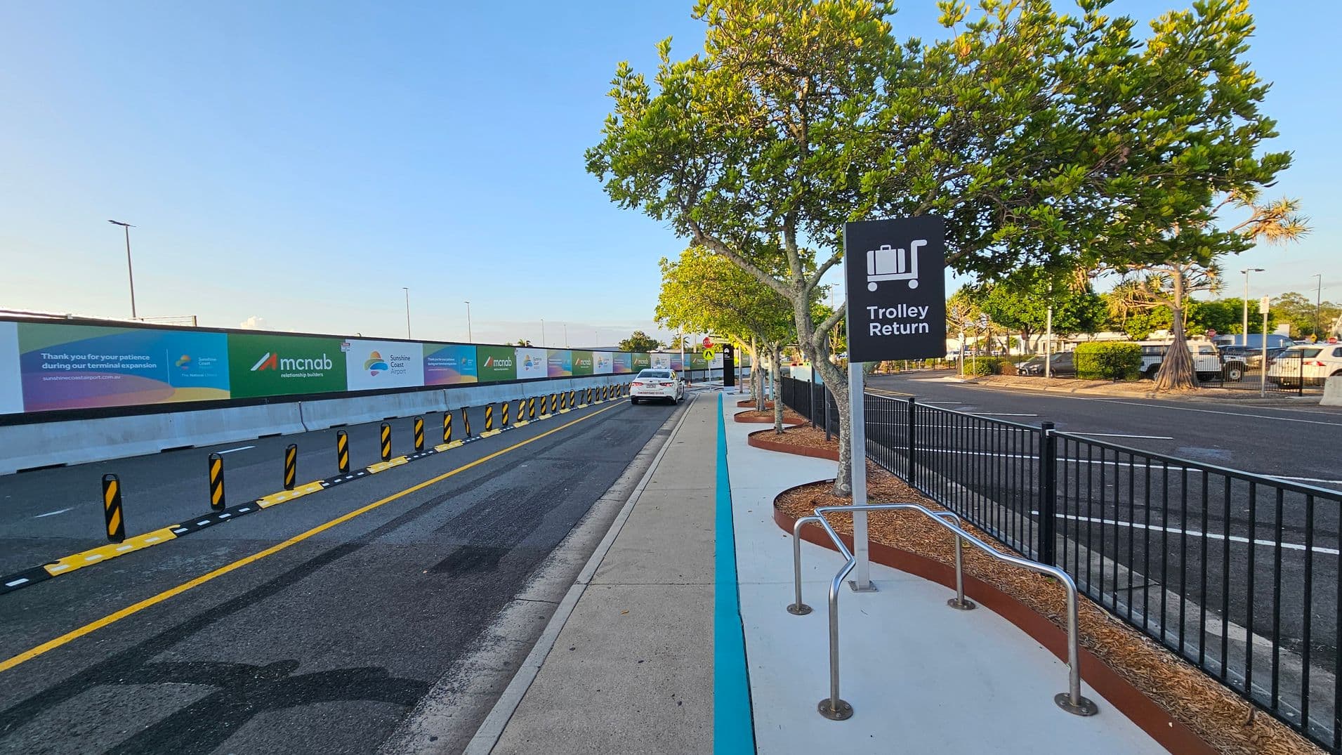 A road with a designated lane on the left, marked by yellow lines and black-and-yellow bollards. A white car is parked further down the lane. On the right, a pavement with a "Trolley Return" sign and metal trolley racks. Trees with green foliage line the pavement, and a black metal fence separates the pavement from a car park. Advertising banners are displayed on a wall to the left. The ground is asphalt and concrete, with mulch around the tree bases.