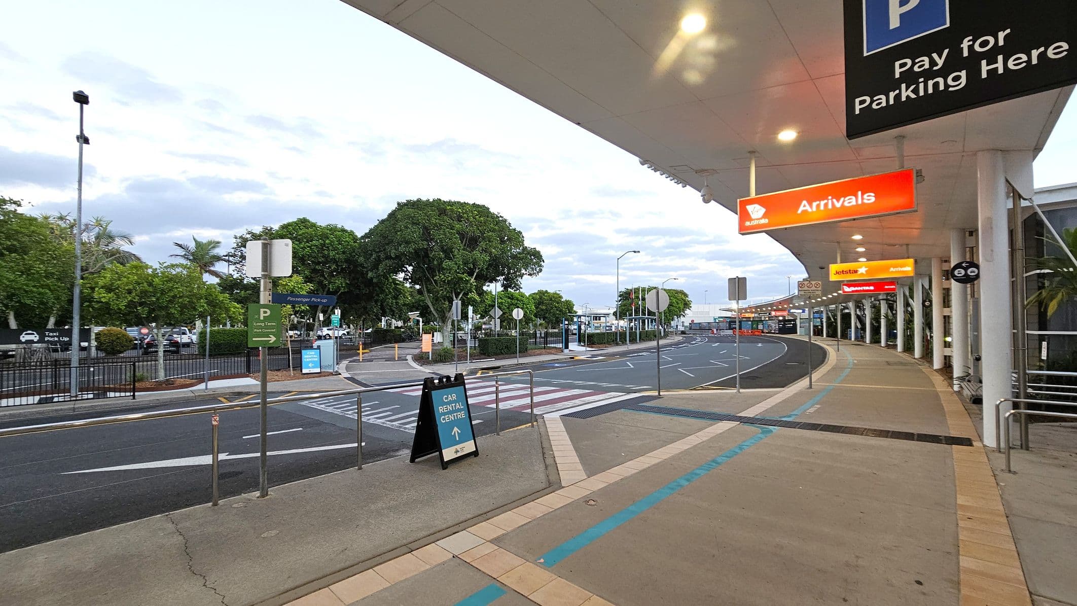 A covered walkway at an airport with a beige tiled floor and a white ceiling with lights. On the right, there are signs for "Arrivals" in orange and "Pay for Parking Here" in blue. The road curves to the left with marked lanes and pedestrian crossings. On the left, there are trees and signs for "Passenger Pick-up" and "Car Rental Centre". A fence separates the walkway from the road.
