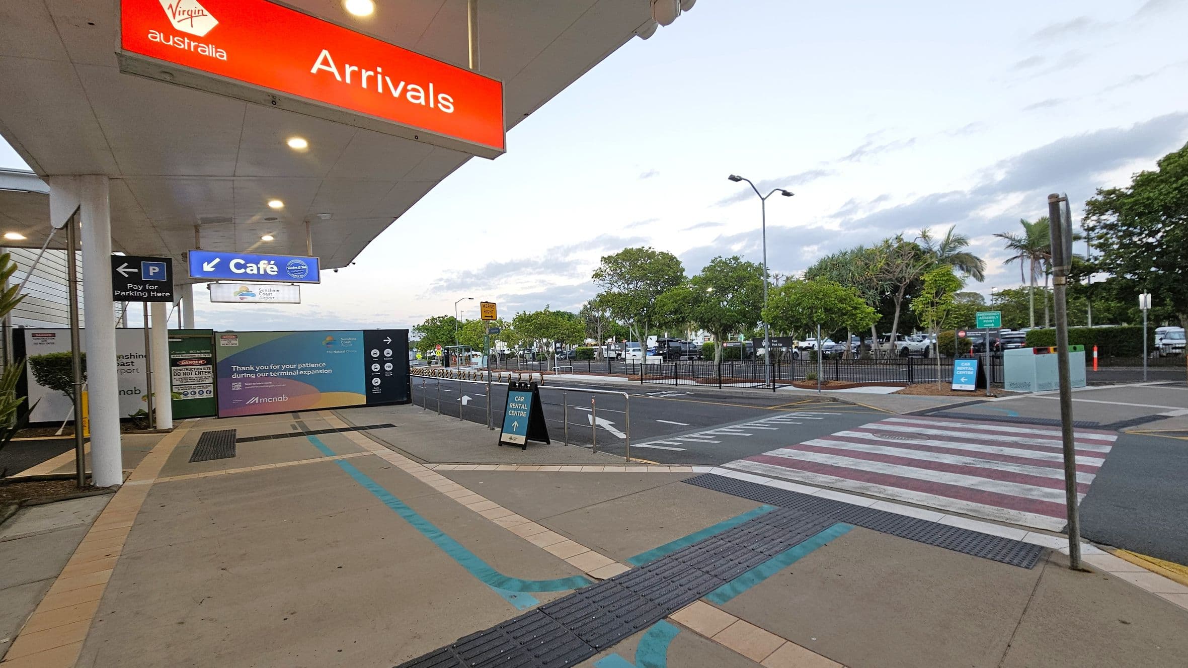 Under a covered walkway, a red sign reads "Arrivals" with "Virgin Australia" above it. To the left, signs indicate parking payment and a café. A construction barrier displays a message about terminal expansion. In the background, a car park is visible with trees and a few parked cars. A pedestrian crossing with white and purple stripes leads across the road. The pavement is light-coloured with blue and purple tactile paths.