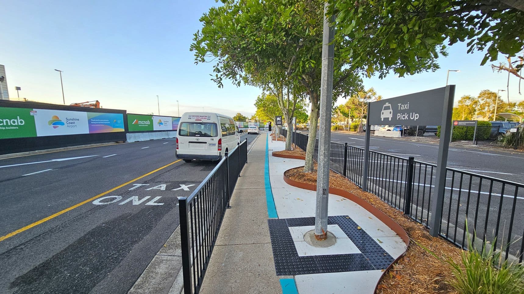 A paved walkway runs through the centre, bordered by black metal fencing on both sides. On the left, a white van is parked in a lane marked "TAXI ONLY," with construction signs along the fence. On the right, a "Taxi Pick Up" sign is visible, with trees planted in mulched beds along the path. The background features more vehicles and a parking area. The ground is concrete with a blue line, and the area is well-lit with street lamps.
