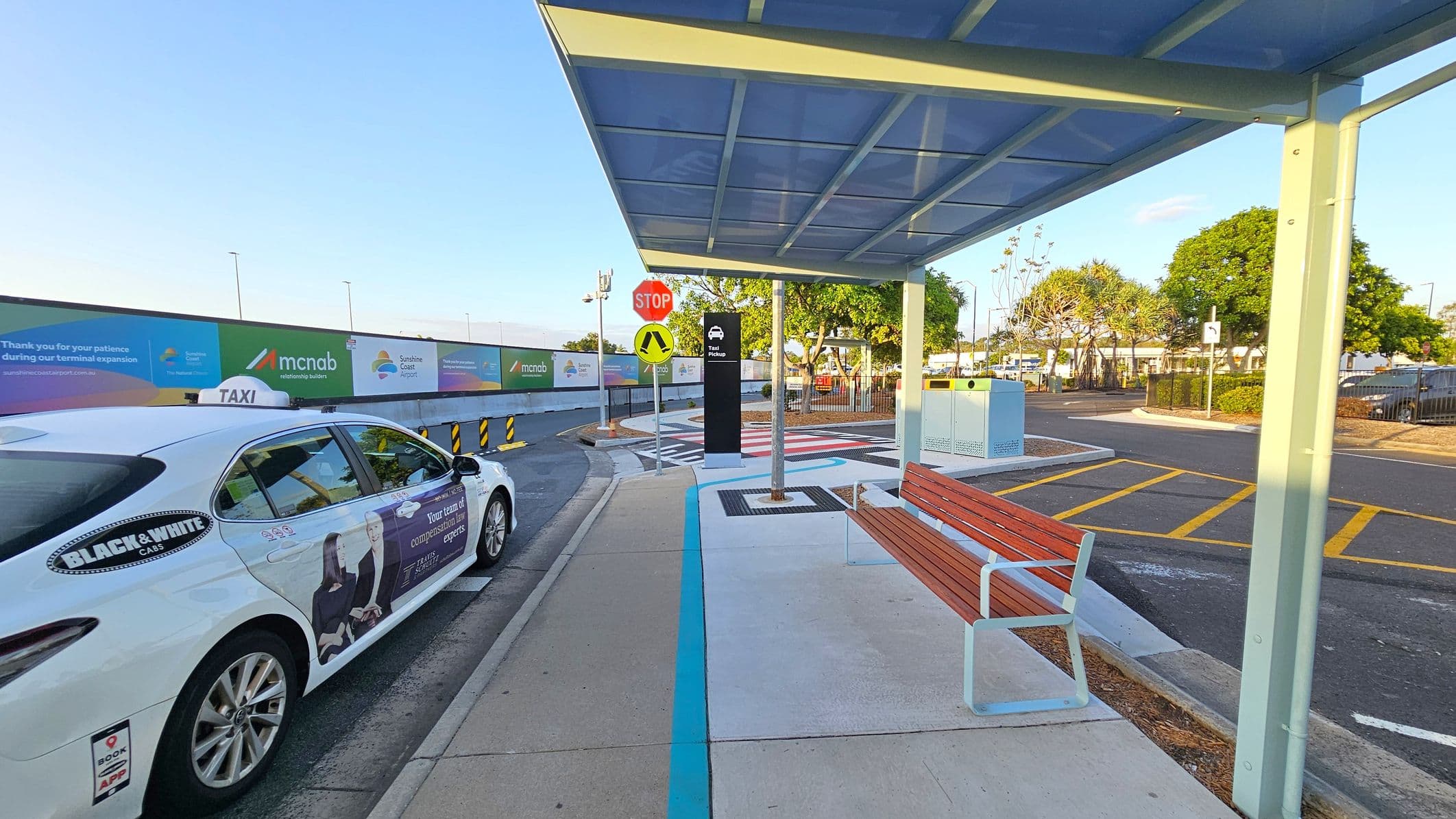 A taxi is parked on the left side of a covered taxi rank area. The shelter has a transparent roof and a wooden bench underneath. A stop sign and pedestrian crossing sign are visible near the centre. In the background, there are trees and a fence, with a road marked with yellow lines. Advertising banners are displayed along a construction fence on the left. The ground is a mix of concrete and asphalt.