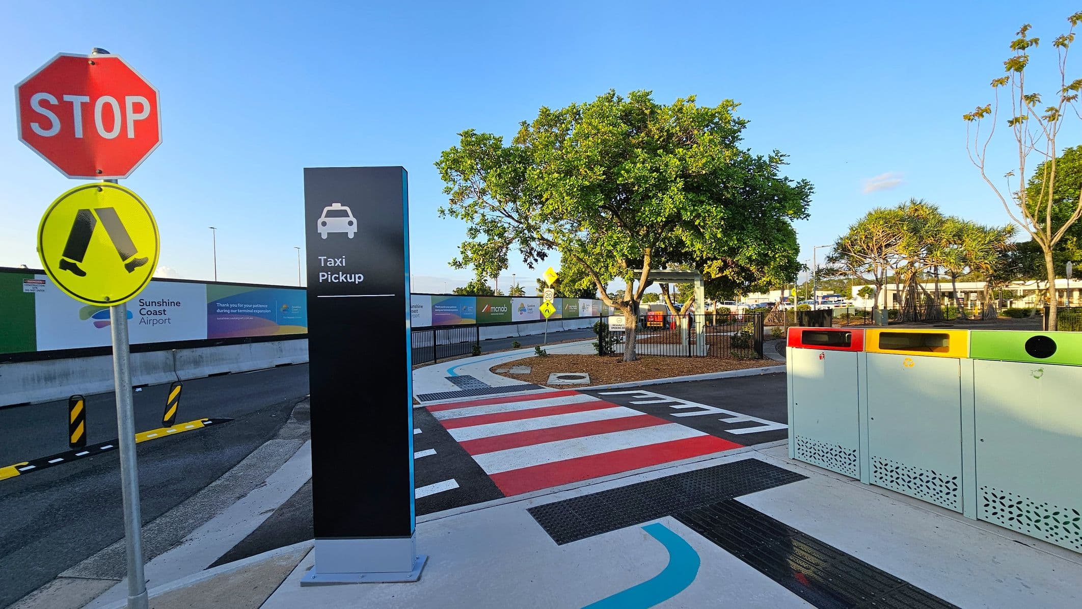 A taxi pickup area with a prominent black sign featuring a taxi icon and the words "Taxi Pickup" in the centre. On the left, a red stop sign and a yellow pedestrian crossing sign are visible. The ground has a red and white striped pedestrian crossing. On the right, there are three waste bins in red, yellow, and green. Trees and a small landscaped area are in the background, with a fence and airport signage visible along the perimeter.