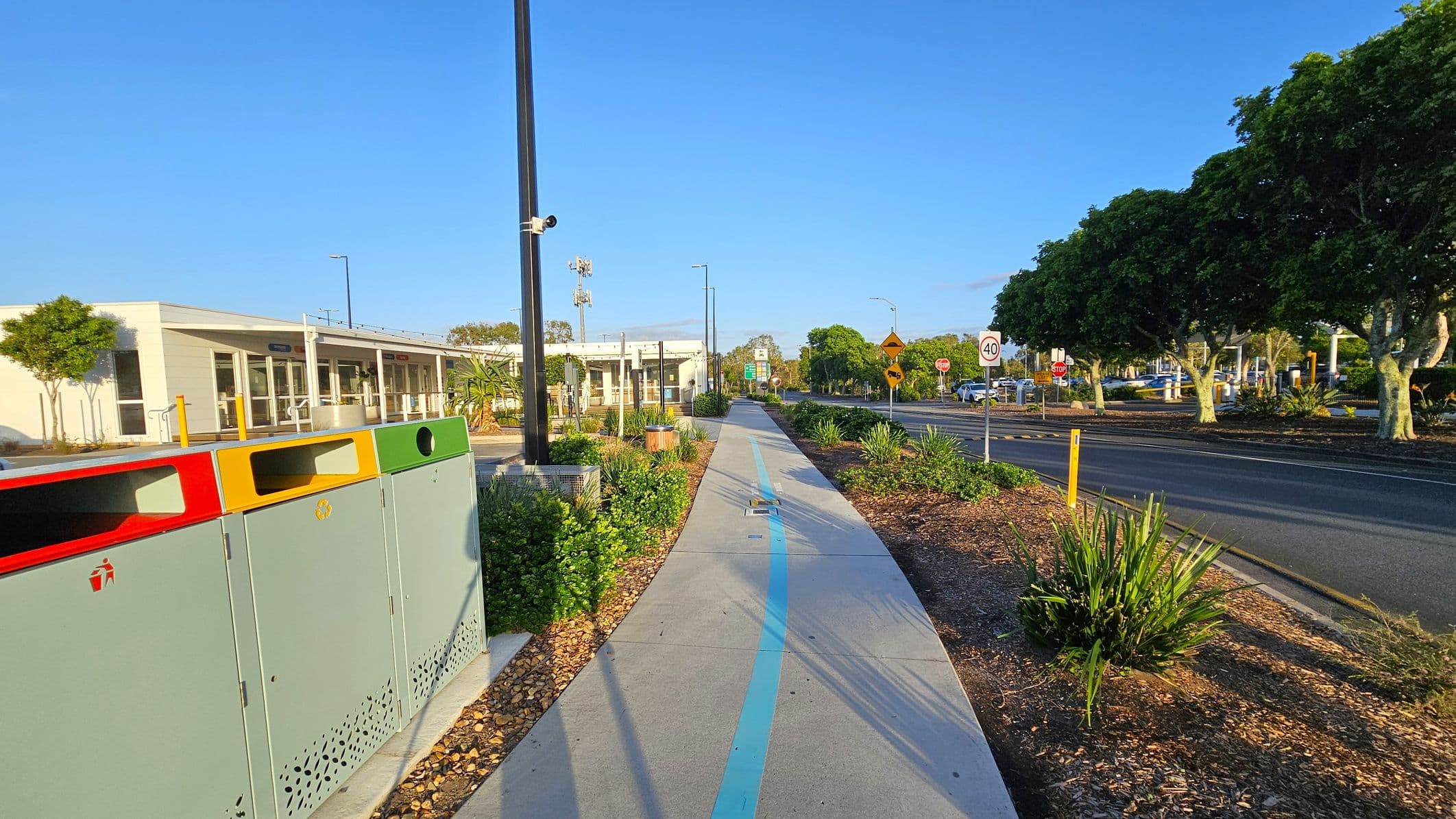 A pathway runs through the centre, marked with a blue line. On the left, there are colourful bins and a modern building with large windows. On the right, a road with a 40 km/h speed limit sign is lined with lush green trees. The foreground features landscaped plants and a tall streetlight. The background shows more trees and a clear sky.