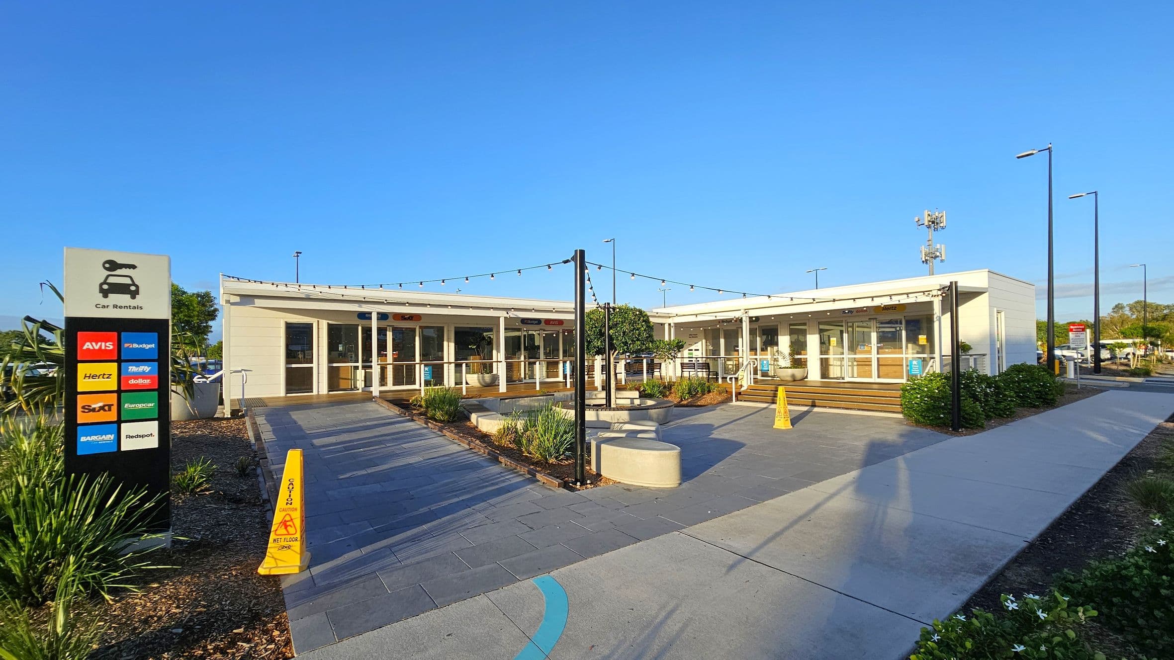 A car rental facility with a modern design. On the left, a sign displays various car rental company logos. The centre features a walkway leading to the entrance, flanked by small plants and a yellow caution sign. The building has large glass windows and a flat roof adorned with string lights. On the right, a paved path runs alongside the building, bordered by greenery. The area is well-lit with tall street lamps.