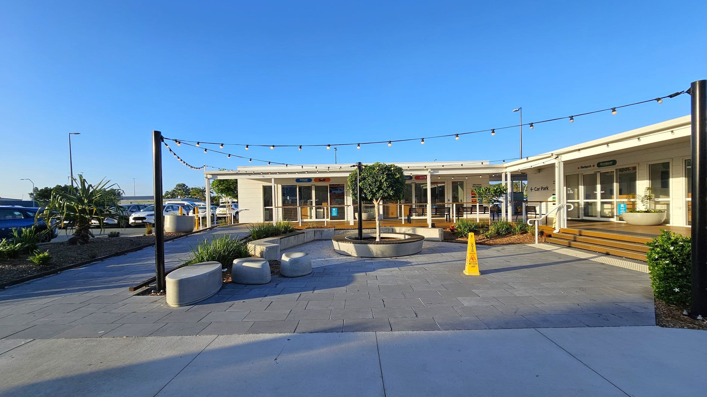 A courtyard with a paved area featuring a circular planter with a small tree in the centre. Surrounding the planter are concrete benches. Overhead, string lights are suspended between black poles. To the right, a building with glass doors and steps leads to a car park. A yellow caution sign is visible near the steps. On the left, there are parked cars and some greenery, including a large plant. The sky is clear and blue.