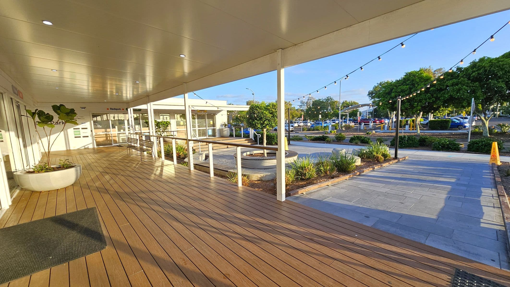 A covered wooden deck with a white ceiling and recessed lights on the left. A large potted plant sits near the entrance of a building with glass doors. A bench is positioned along the railing. On the right, a paved area with string lights overhead leads to a landscaped garden with small trees and shrubs. In the background, a car park with several vehicles and more trees is visible. A yellow caution sign is placed on the pavement.