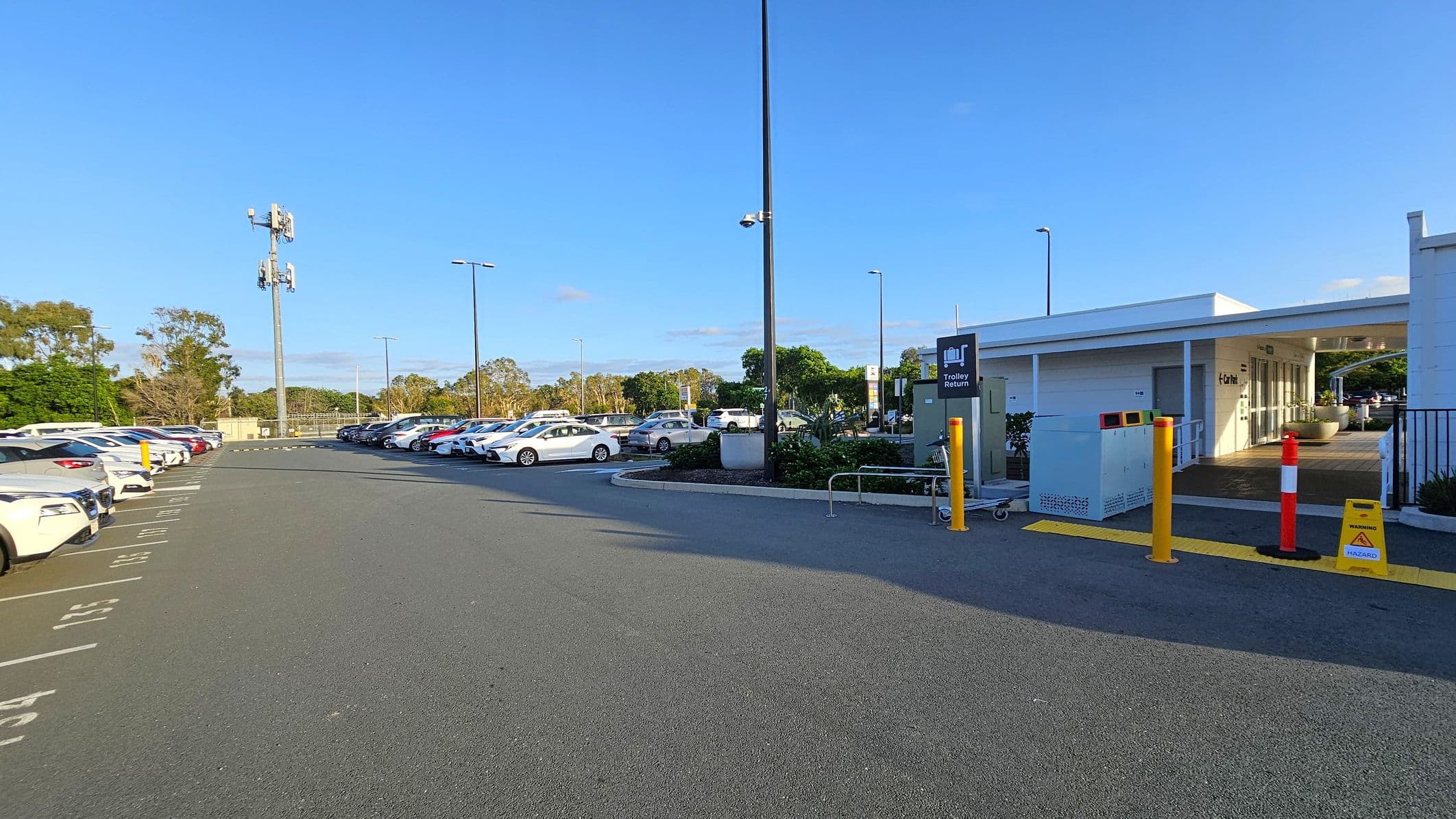 A car park with rows of parked cars on both sides. On the right, there's a white building with a covered entrance and a sign for trolley return. Yellow bollards and a caution sign are near the entrance. A tall pole with security cameras and a mobile tower is visible in the background. The ground is paved, and there are trees and greenery in the distance.