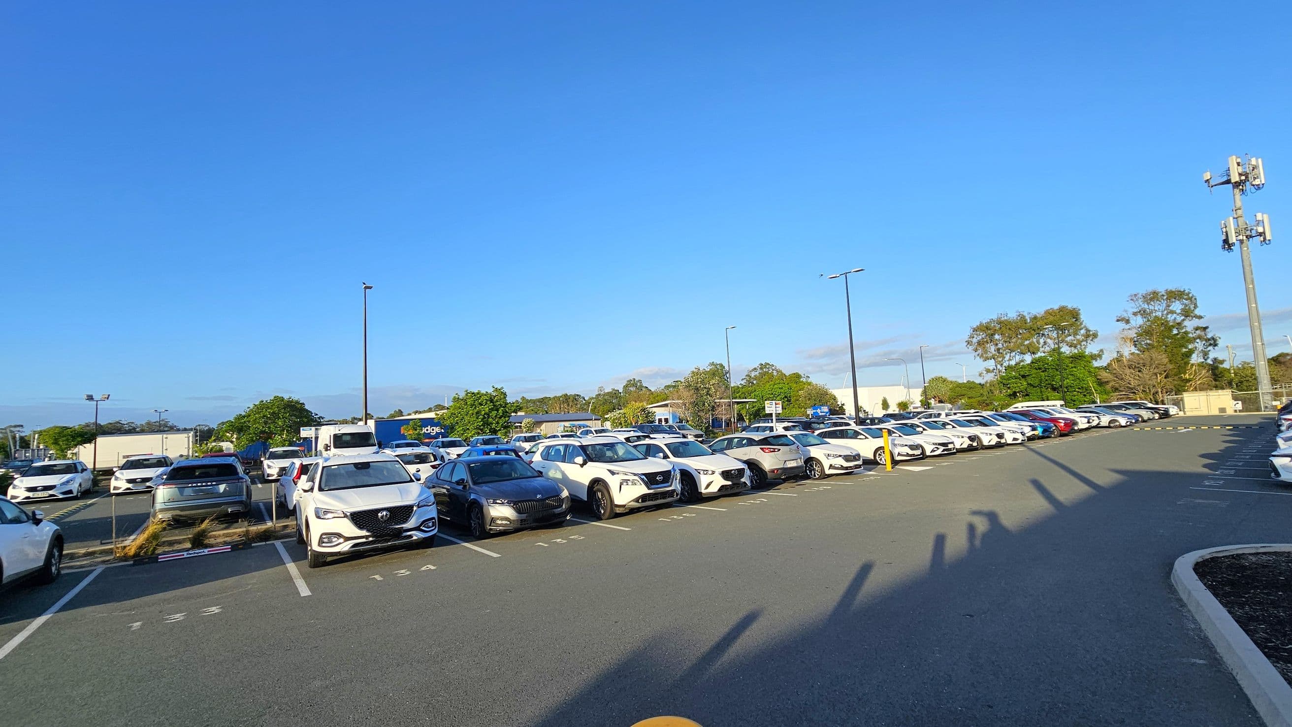 A large car park with multiple rows of parked vehicles, mostly white, stretching from the left to the right. In the background, there are trees and a few buildings. A tall communication tower is visible on the right side. The ground is paved with marked parking spaces, and there are shadows cast by the vehicles and poles. The area is well-lit, suggesting daytime.