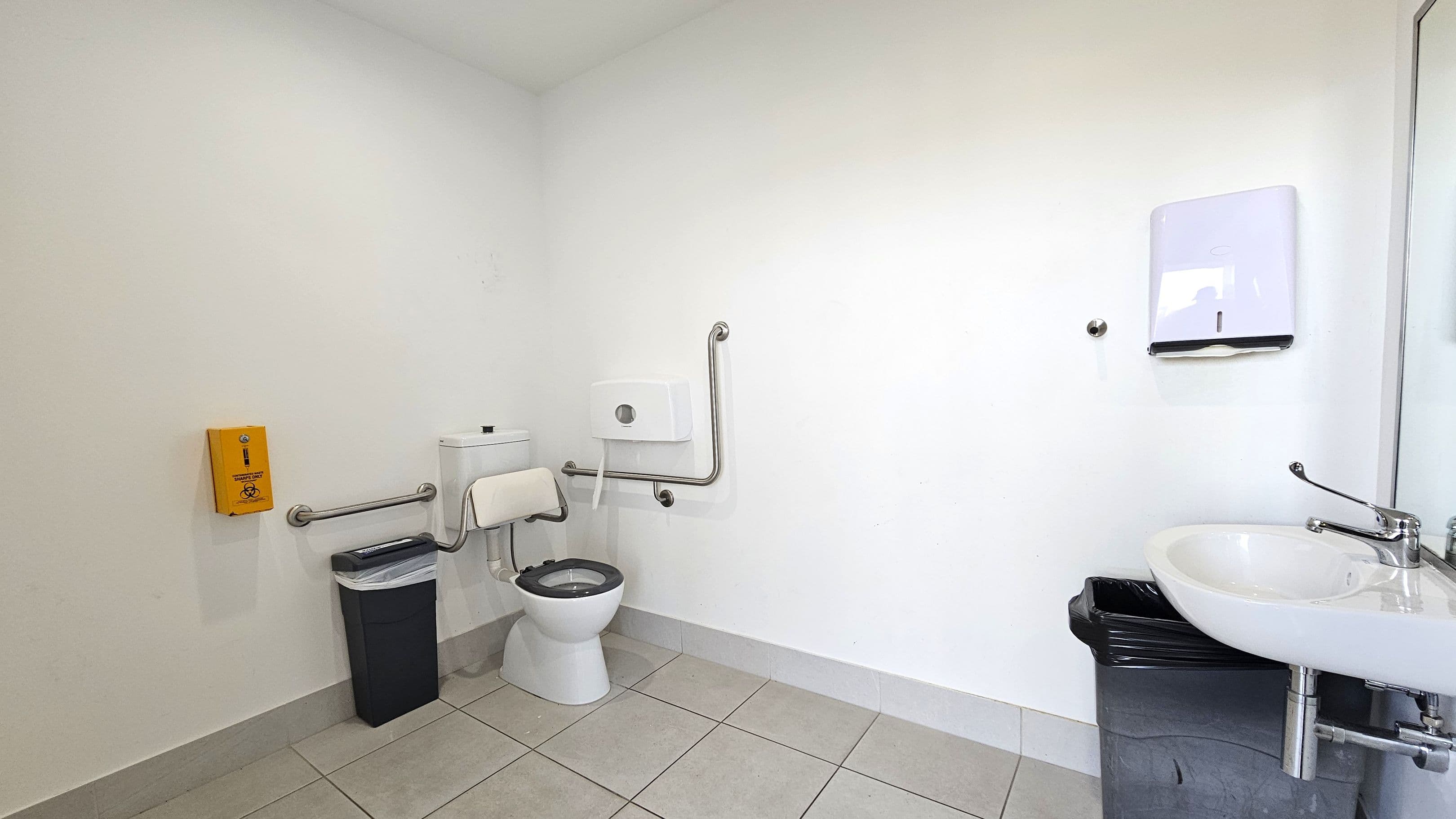 A clean, accessible bathroom with a white toilet featuring support rails on both sides. To the left, a yellow emergency assistance box is mounted on the wall above a black bin. The floor is tiled in light grey. On the right, a white sink with a lever tap is positioned above another black bin. A paper towel dispenser is mounted on the wall above the sink. The walls are plain white, and the space is well-lit.