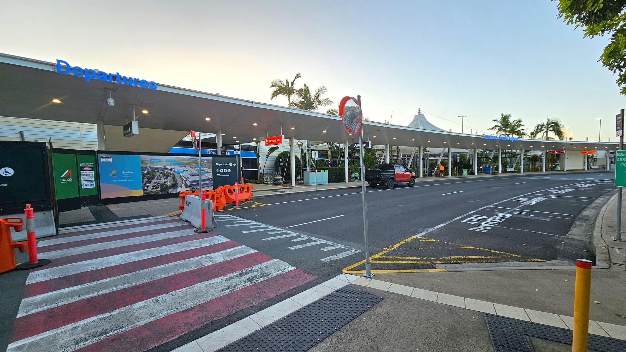 A covered airport departures area with a long, white canopy extending across the scene. On the left, there are construction barriers and signs, including a blue "Departures" sign. In the middle, a pedestrian crossing with red and white stripes leads to the road. On the right, a red vehicle is parked near the kerb. Palm trees are visible in the background, and the road has markings for passenger drop-off. The area is well-lit with overhead lights.