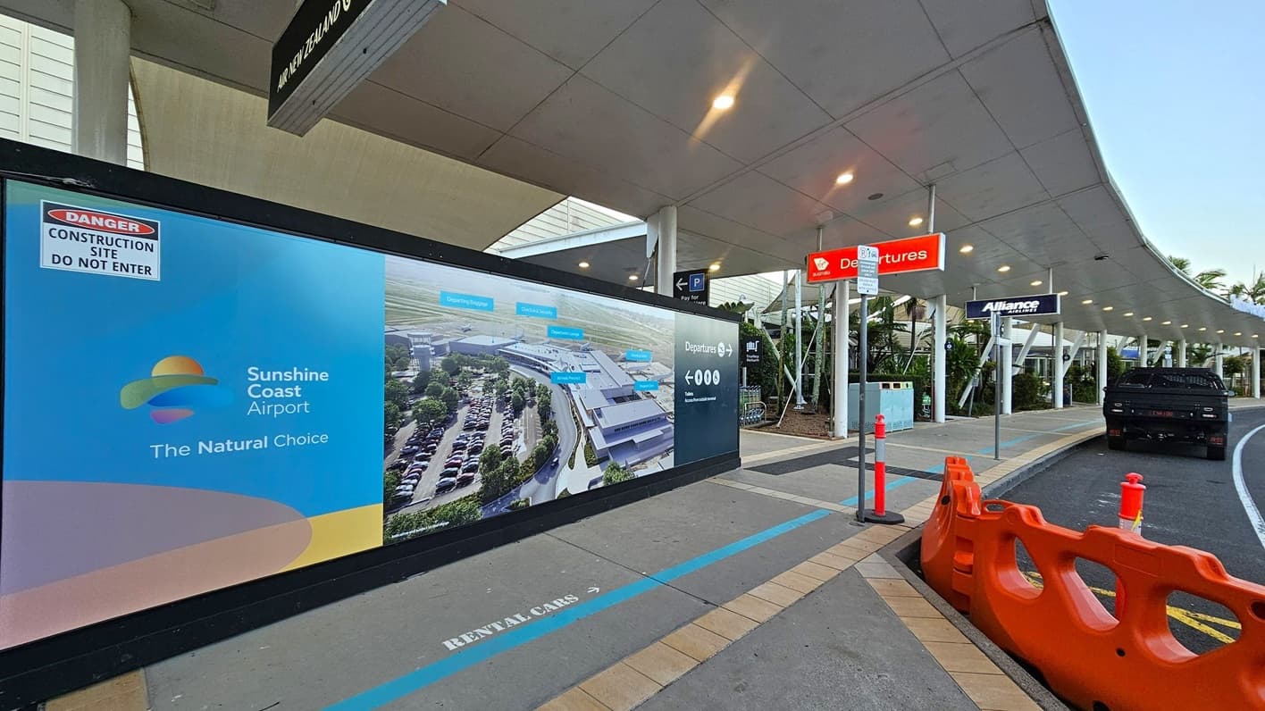 A covered walkway at an airport with a large advertisement on the left for Sunshine Coast Airport, featuring a map and the slogan "The Natural Choice." Above, a sign for Air New Zealand is visible. On the right, a red "Departures" sign hangs above the pavement, where a black vehicle is parked. Orange barriers and traffic cones line the kerb. The ceiling is white with lights, and the floor has a marked path for rental cars. Palm trees are visible in the background.