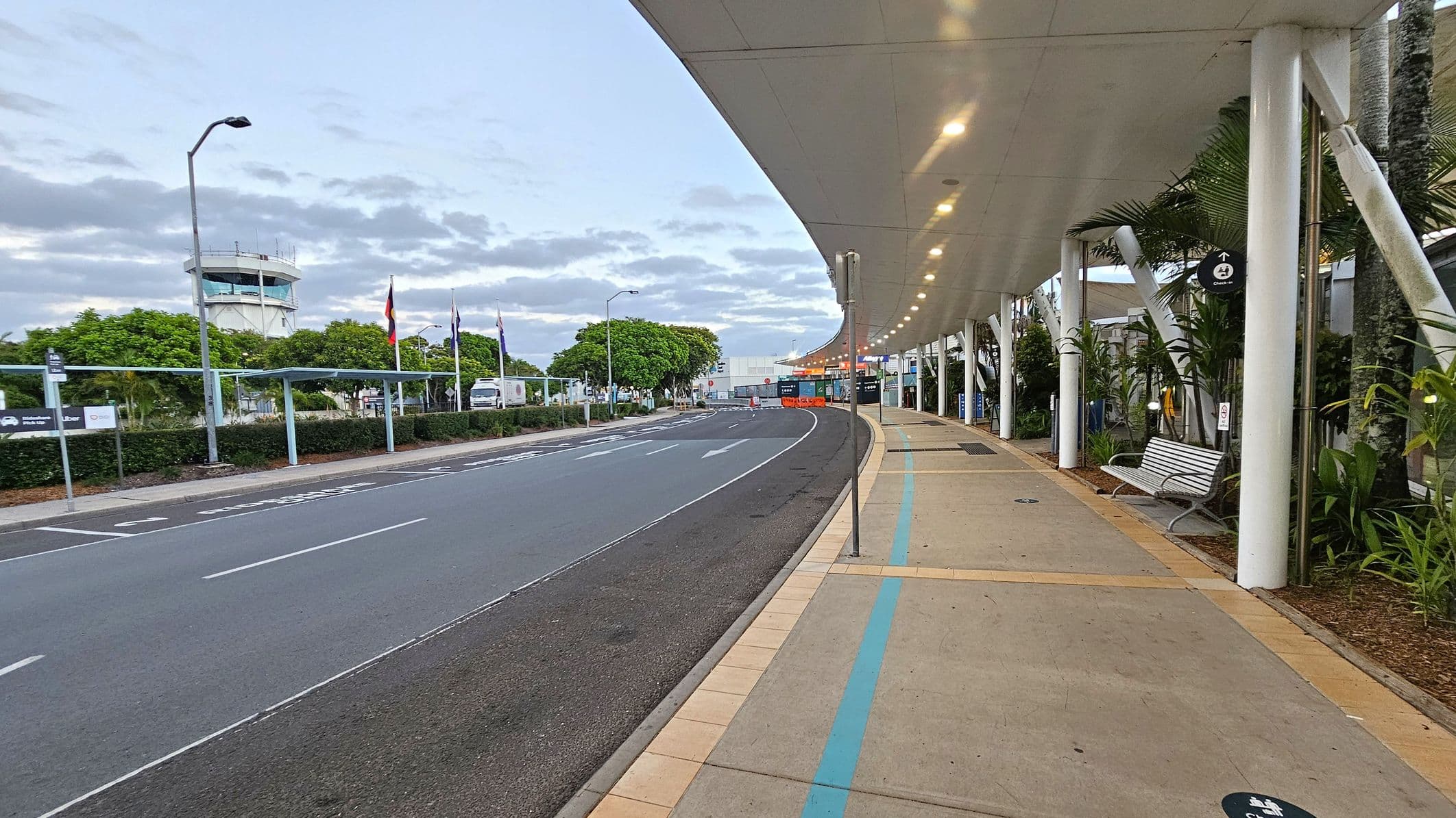 A curved road runs through the centre, with a pedestrian walkway on the right. The walkway is covered by a white ceiling with lights and supported by white pillars. A bench and some plants are along the walkway. On the left, there are trees and several flags near a fence. A control tower is visible in the background. The road has marked lanes and a sign indicating a pick-up area. The scene is well-lit, suggesting early morning or late afternoon.