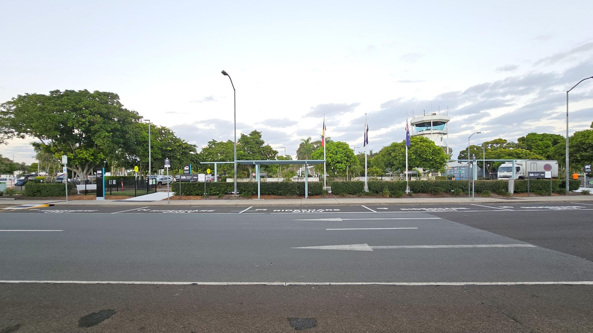 A road with marked parking spaces in the foreground, labelled for rideshare and passenger pick-up. Behind the road, there is a fenced area with a bus shelter and several flagpoles displaying flags. Trees and shrubs are scattered throughout the area. In the background, a control tower is visible, along with a parked truck. Street lamps are positioned along the road and within the fenced area.