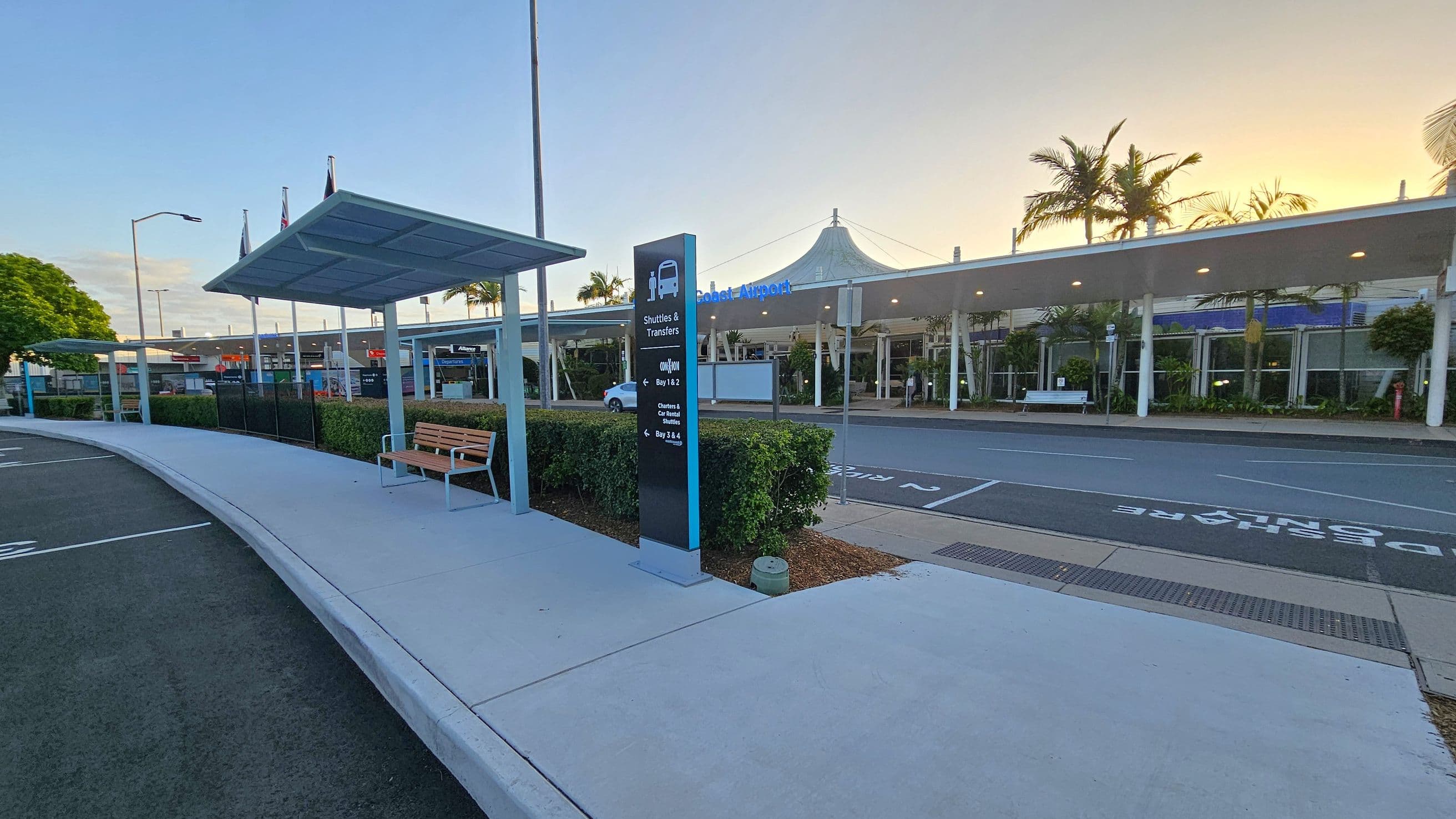 A covered walkway with a bench on the left, next to a sign for shuttles and transfers. The pavement is light grey, and the road is adjacent. In the background, a building with a canopy and glass windows is visible, with palm trees and flags on poles. The sky is a gradient of light colours, suggesting early morning or late afternoon.