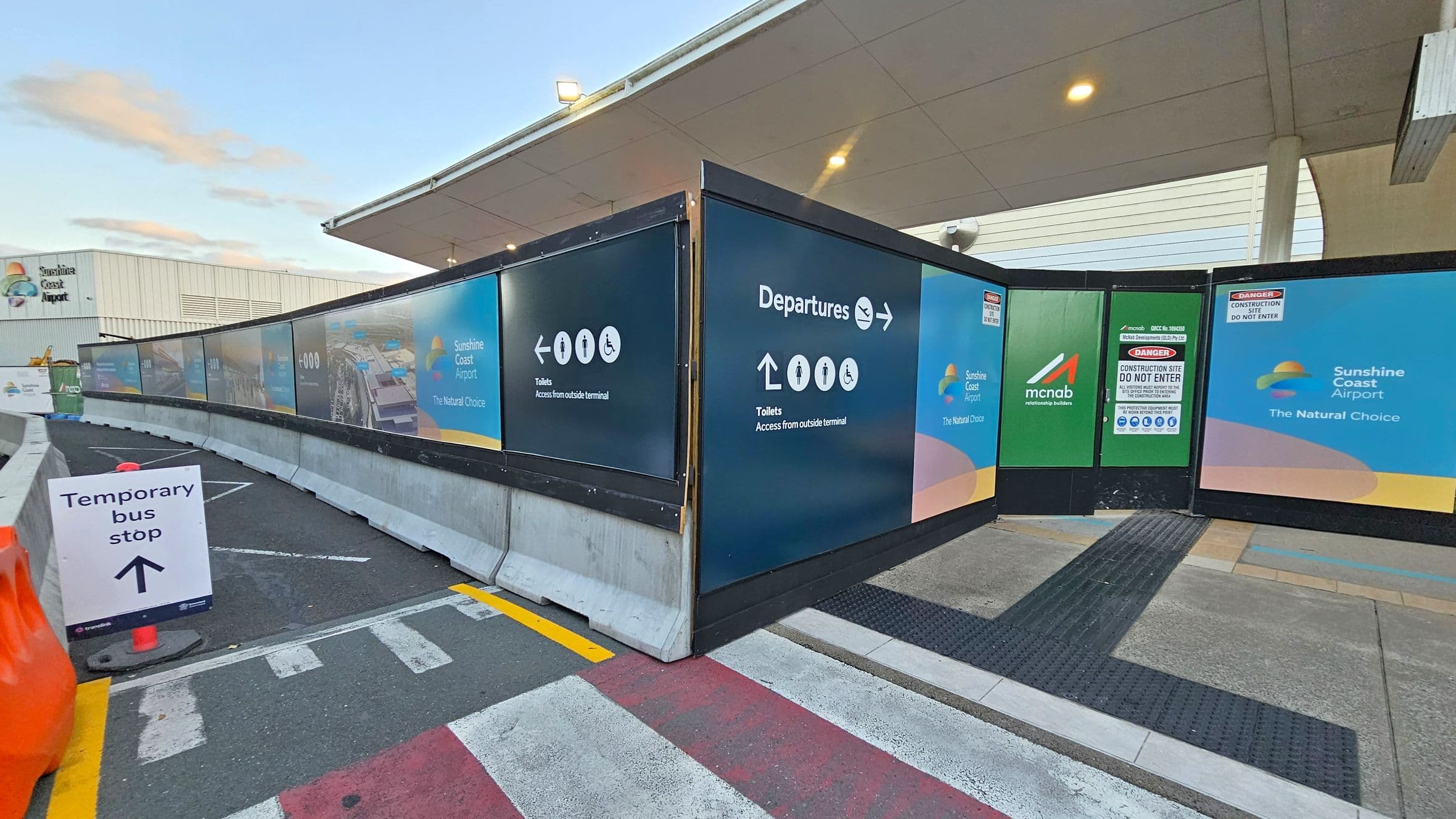 Construction barriers with signage for Sunshine Coast Airport are on the right, displaying directions to departures and toilets. A green panel with "mcnab" and "Do Not Enter" warnings is visible. On the left, a temporary bus stop sign is placed on the ground near orange traffic cones. The ground features a mix of grey, red, and yellow markings. The background shows a building with the Sunshine Coast Airport logo and a partially visible roof structure.