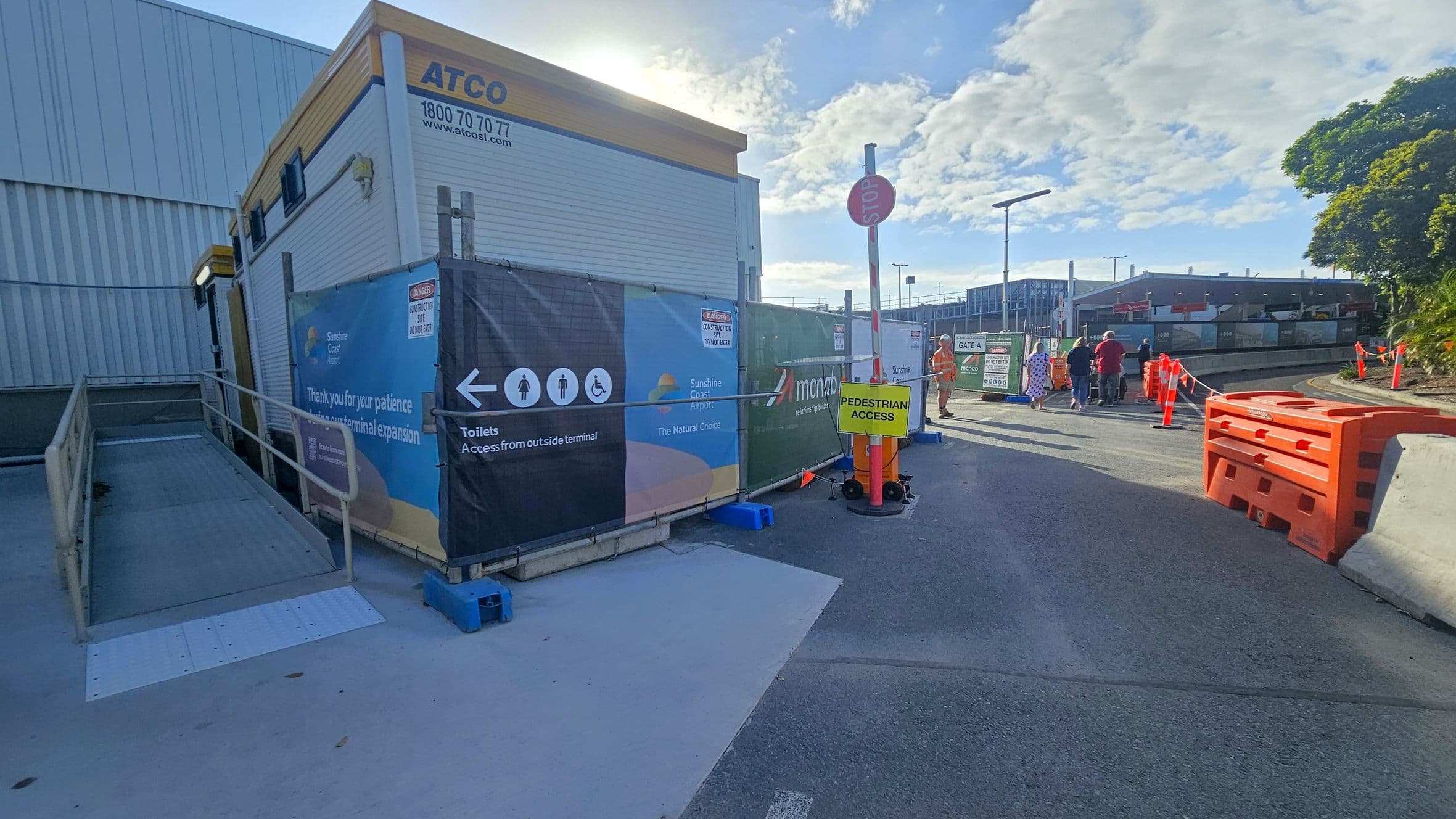 A construction area with a portable building on the left, marked with "ATCO" and contact details. A ramp leads up to the building. A fence with signage directs to toilets and indicates construction. In the centre, a "Pedestrian Access" sign is attached to a pole. On the right, people walk along a path bordered by orange barriers and cones. A tree and industrial buildings are visible in the background. The ground is paved, and the sky is partly cloudy.