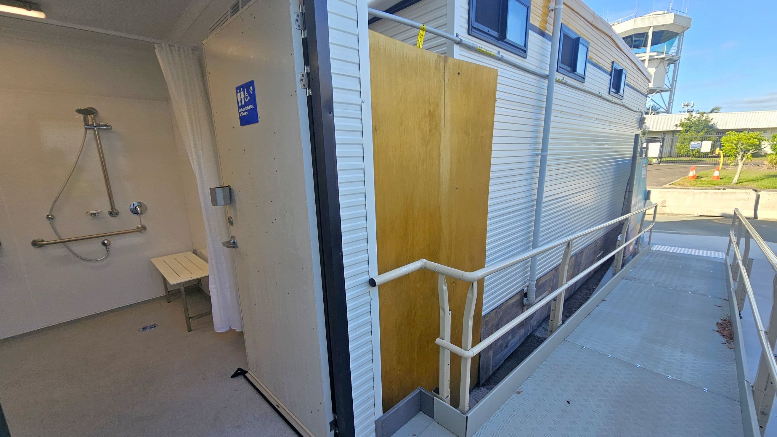 A shower room is visible on the left, featuring a showerhead, grab bars, and a foldable bench. The door is open, displaying a sign indicating accessibility. On the right, a metal ramp with railings leads to the entrance of a portable building with small windows. In the background, there is a control tower and some greenery, along with traffic cones on the pavement.