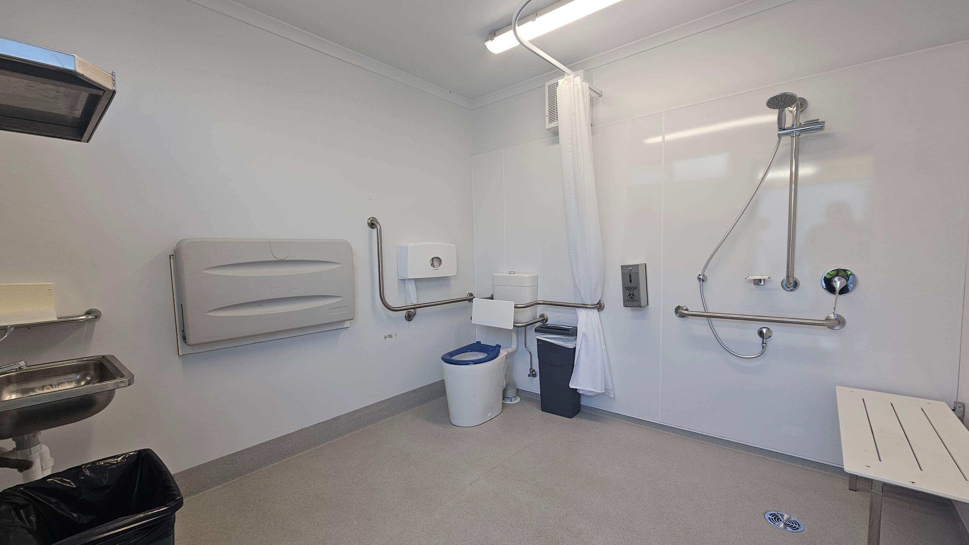 A spacious, accessible bathroom with a grey floor and white walls. On the left, a stainless steel sink with a soap dispenser above and a black bin below. In the centre, a toilet with a blue seat, surrounded by metal grab bars, a paper dispenser, and a bin. On the right, a shower area with a handheld showerhead, grab bars, and a white curtain. A foldable bench is attached to the wall. The ceiling has a fluorescent light fixture.
