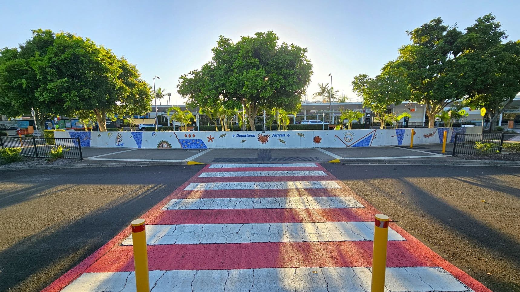 A red and white striped pedestrian crossing leads to a painted wall with colourful designs and arrows indicating "Departures" and "Arrivals". Behind the wall, there are several large green trees and a few smaller palm trees. The ground is asphalt, and there are yellow bollards near the crossing. The area is bordered by black metal fencing on both sides. In the background, there are buildings partially visible through the trees.