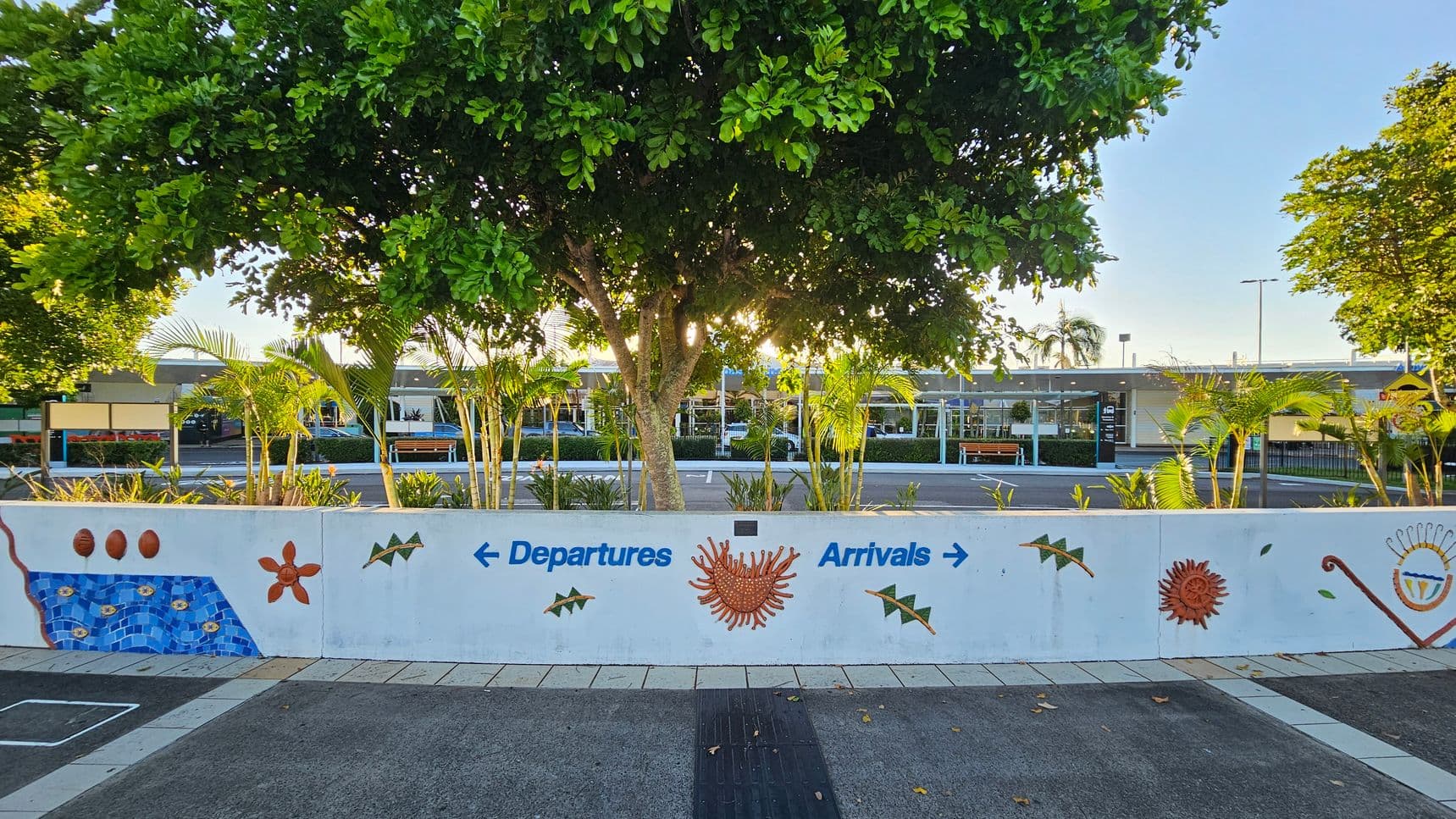 A concrete wall with colourful mosaic designs and the words "Departures" with a left arrow and "Arrivals" with a right arrow. In the foreground, there's a paved walkway with a tactile strip. Behind the wall, lush green trees and small palm plants are visible. In the background, a building with a flat roof and glass windows is partially obscured by the foliage. The sunlight filters through the trees, casting shadows on the ground.