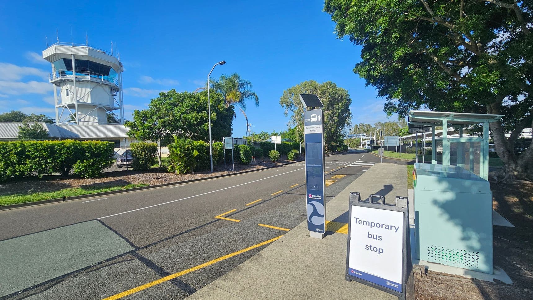 On the left, there is a white control tower with a blue-tinted glass section at the top, surrounded by greenery and a low building with a grey roof. On the right, a temporary bus stop sign is visible next to a small, light blue shelter. The road runs through the centre, with yellow markings and a streetlamp. Trees with dense foliage are on the right, providing shade. The pavement is grey, and the sky is clear and blue.