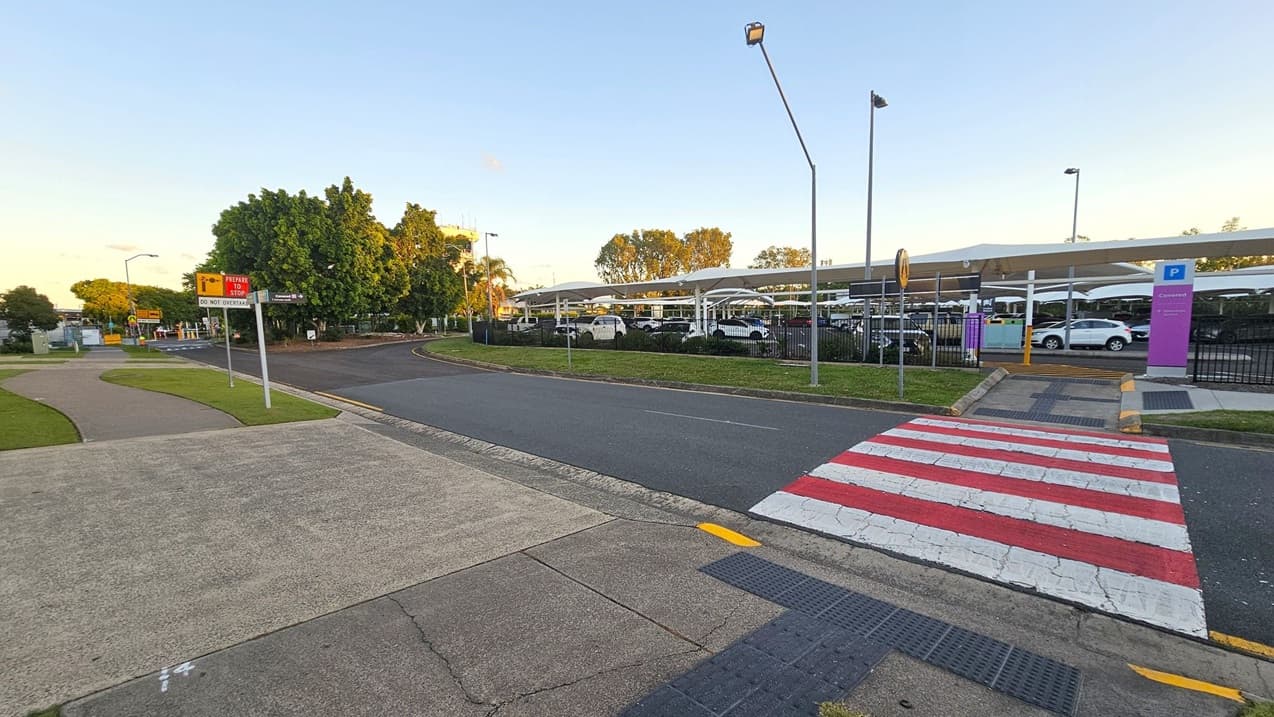 A road with a red and white striped pedestrian crossing in the foreground. To the left, there's a footpath with grass and a sign that reads "PEAKER STOP DO NOT OVERTAKE." In the background, a parking area with several cars is visible, covered by a white canopy. Trees and streetlights are scattered throughout the scene, and a purple sign indicating "Covered" parking is on the right. The sky is clear, and the area is well-lit.