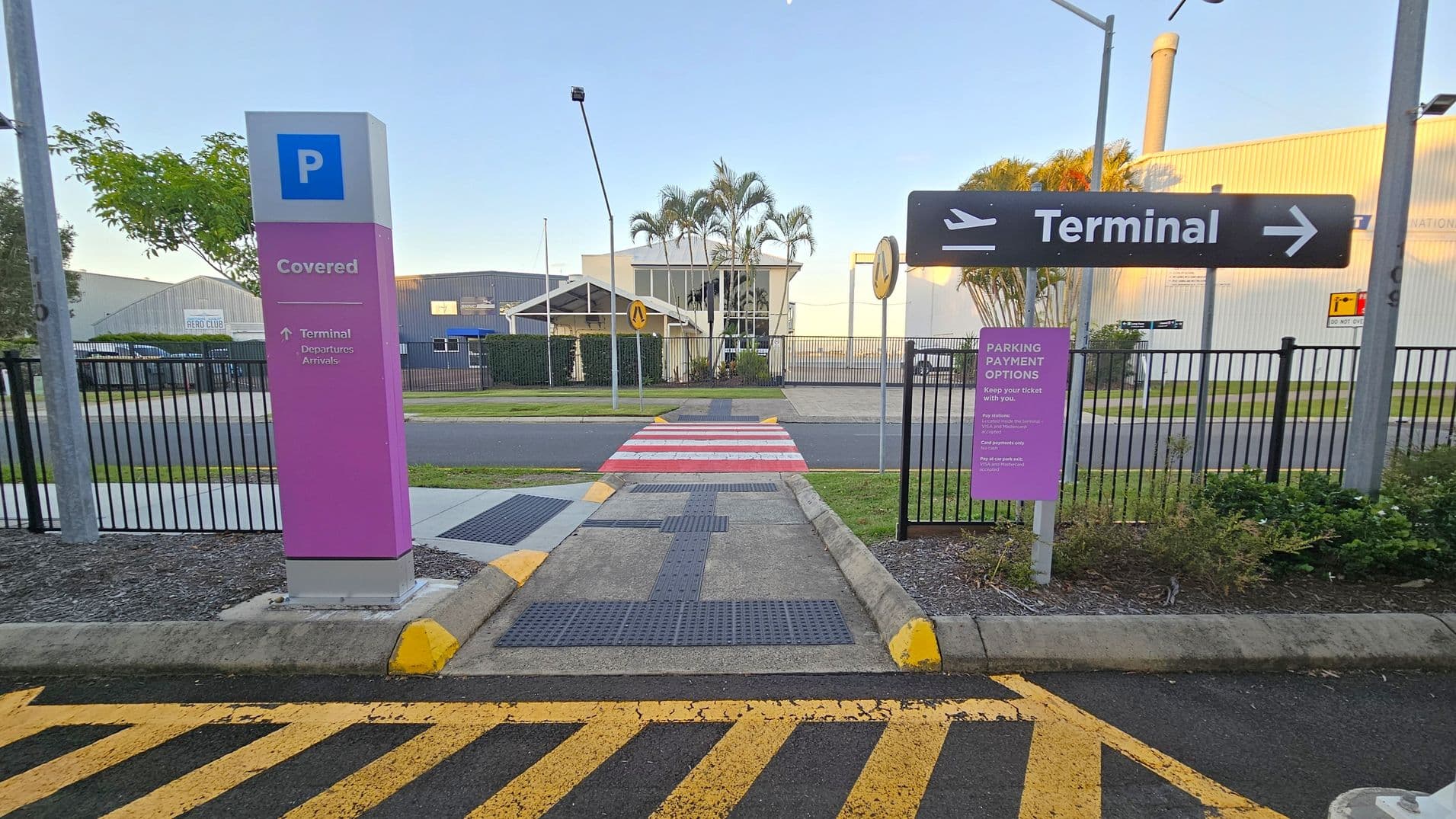 A pathway leads to a zebra crossing with red and white stripes. On the left, a pink sign indicates covered parking and directions to the terminal. On the right, a sign points to the terminal with an aeroplane icon. A purple sign provides parking payment options. The background features a building with palm trees and a fence. The ground has yellow and black striped markings, and there are yellow kerb stops.