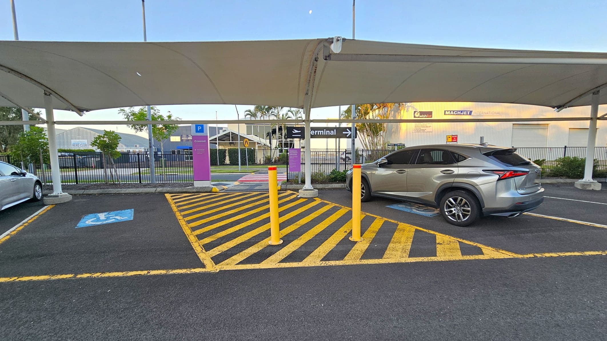 A covered parking area with a silver SUV parked on the right in a space marked with a blue disabled parking symbol. Yellow and black diagonal lines are painted on the ground in the centre, with three yellow bollards. A signpost with "P Covered" is visible. In the background, there are signs for "Terminal" and "Machjet International" on a building. The area is enclosed by a black metal fence, and there are some trees and plants near the fence. The ceiling is a white canopy structure.