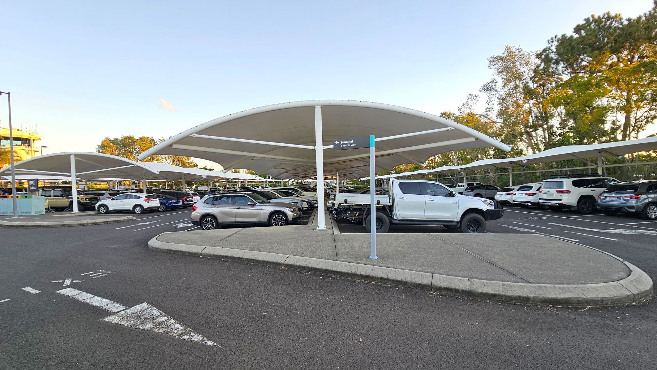 A car park with a covered structure featuring white, curved canopies supported by poles. Numerous vehicles, including a white pickup truck and various cars, are parked beneath the shelters. The ground is paved with asphalt, and a white arrow is painted on the surface, indicating direction. A signpost points towards a terminal, suggesting a nearby airport or transport hub. Trees with green foliage are visible in the background.