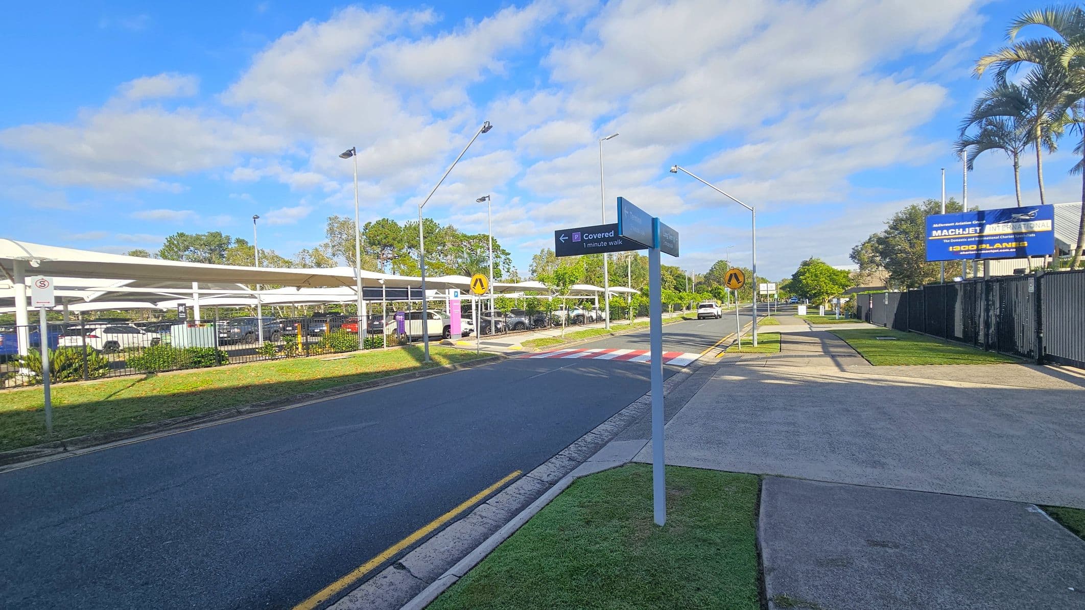 A road with a pedestrian crossing in the centre, flanked by a grassy verge on the left and a pavement on the right. On the left, there is a covered parking area with several parked cars and a fence. A signpost indicates directions. On the right, a large billboard is visible above a fence, with palm trees and other greenery nearby. The sky is partly cloudy.