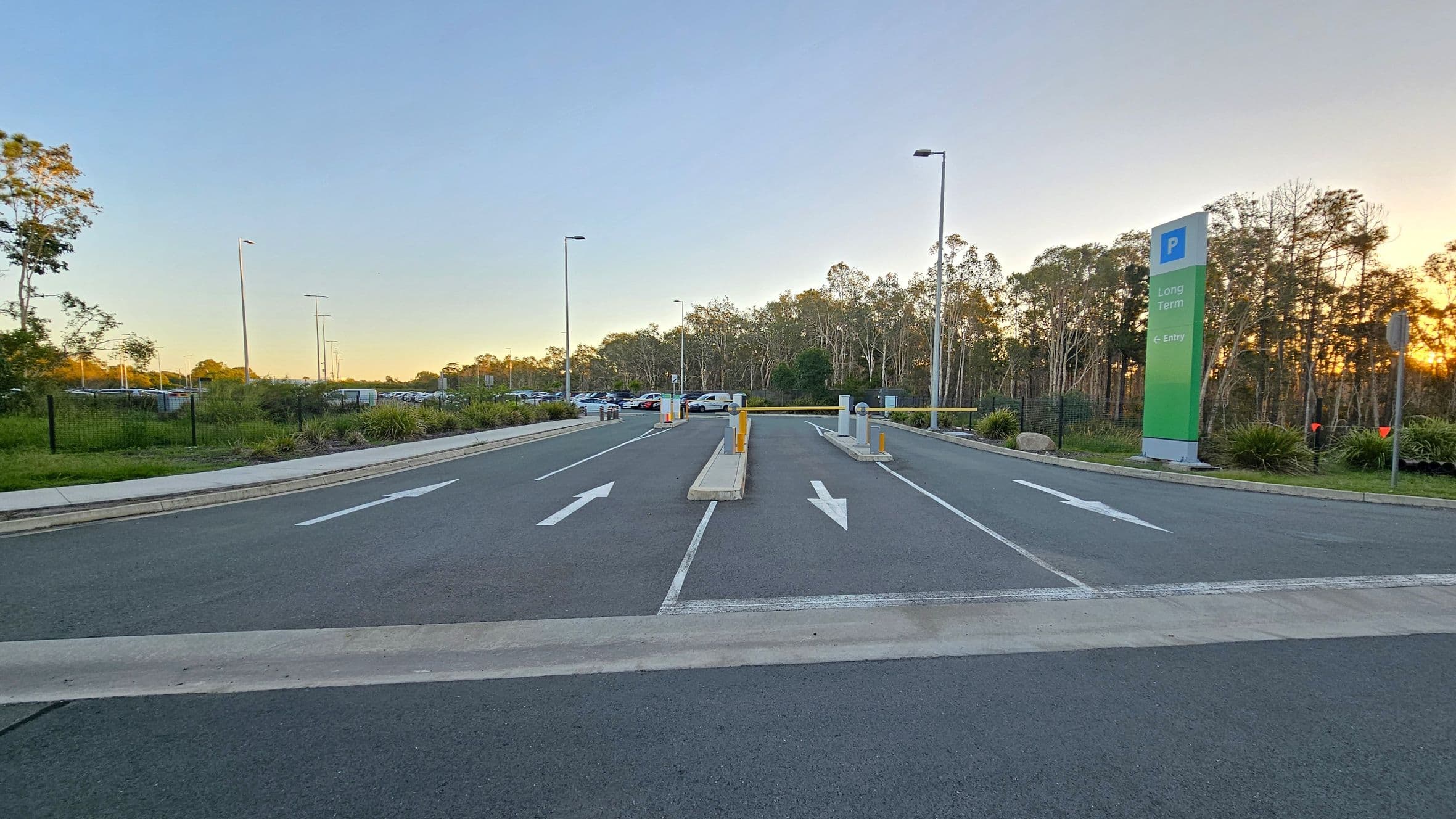 A road with two lanes marked by white arrows pointing forward. In the middle, there are barriers and bollards. On the right, a tall sign reads "Long Term" and "E-Entry" with a parking symbol. The left side has grassy areas and trees, with a row of parked cars visible in the distance. The background features a line of trees, and the lighting suggests a sunset. Streetlights are positioned along the road.