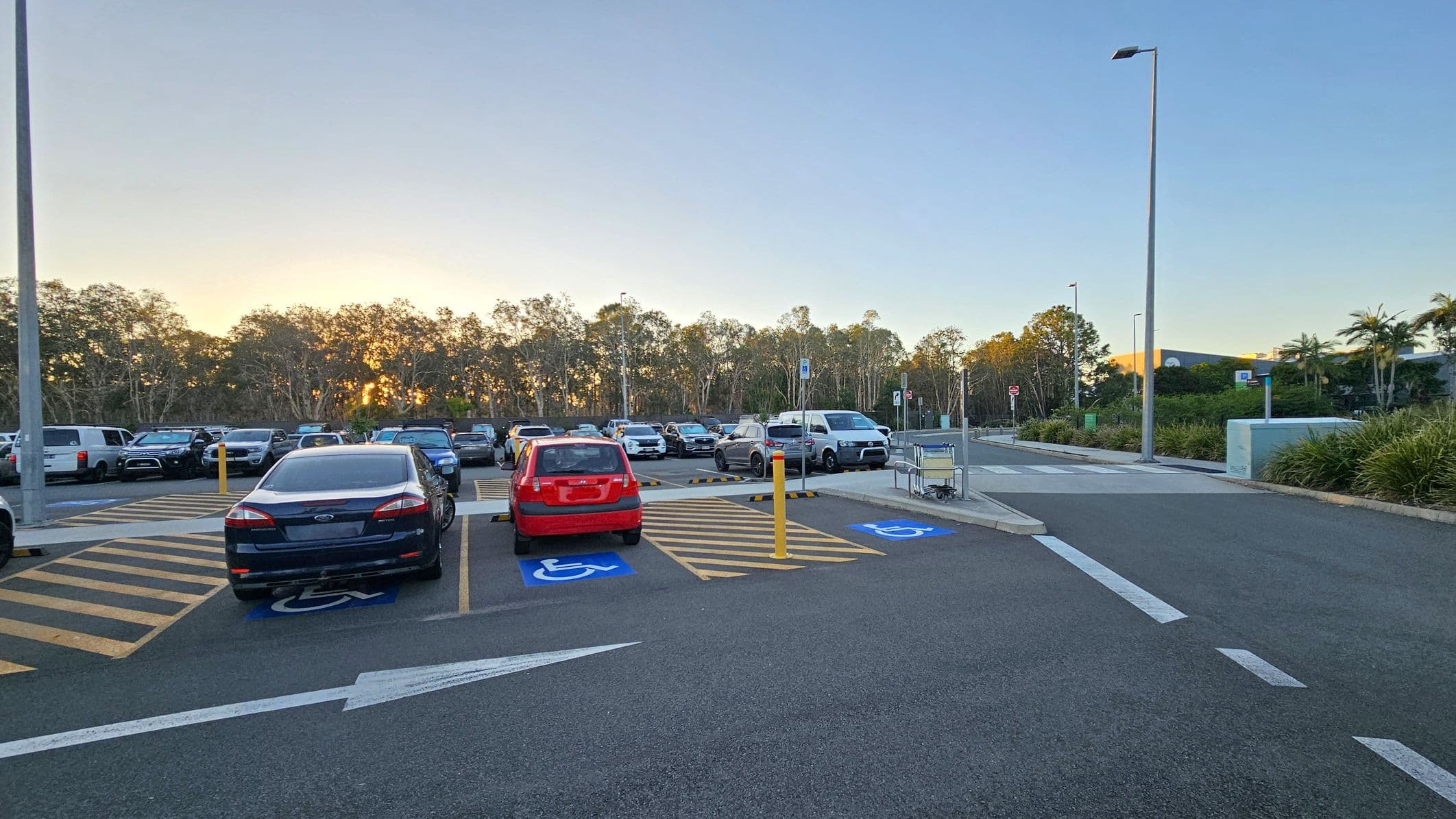 A car park with several vehicles, including a red car and a dark blue car, parked in designated disabled spaces marked with blue paint and wheelchair symbols. Yellow-striped areas separate the spaces. A shopping trolley is on the right near a pavement. Tall streetlights line the area, and trees are visible in the background. The ground is asphalt, and there are directional arrows painted on the surface.