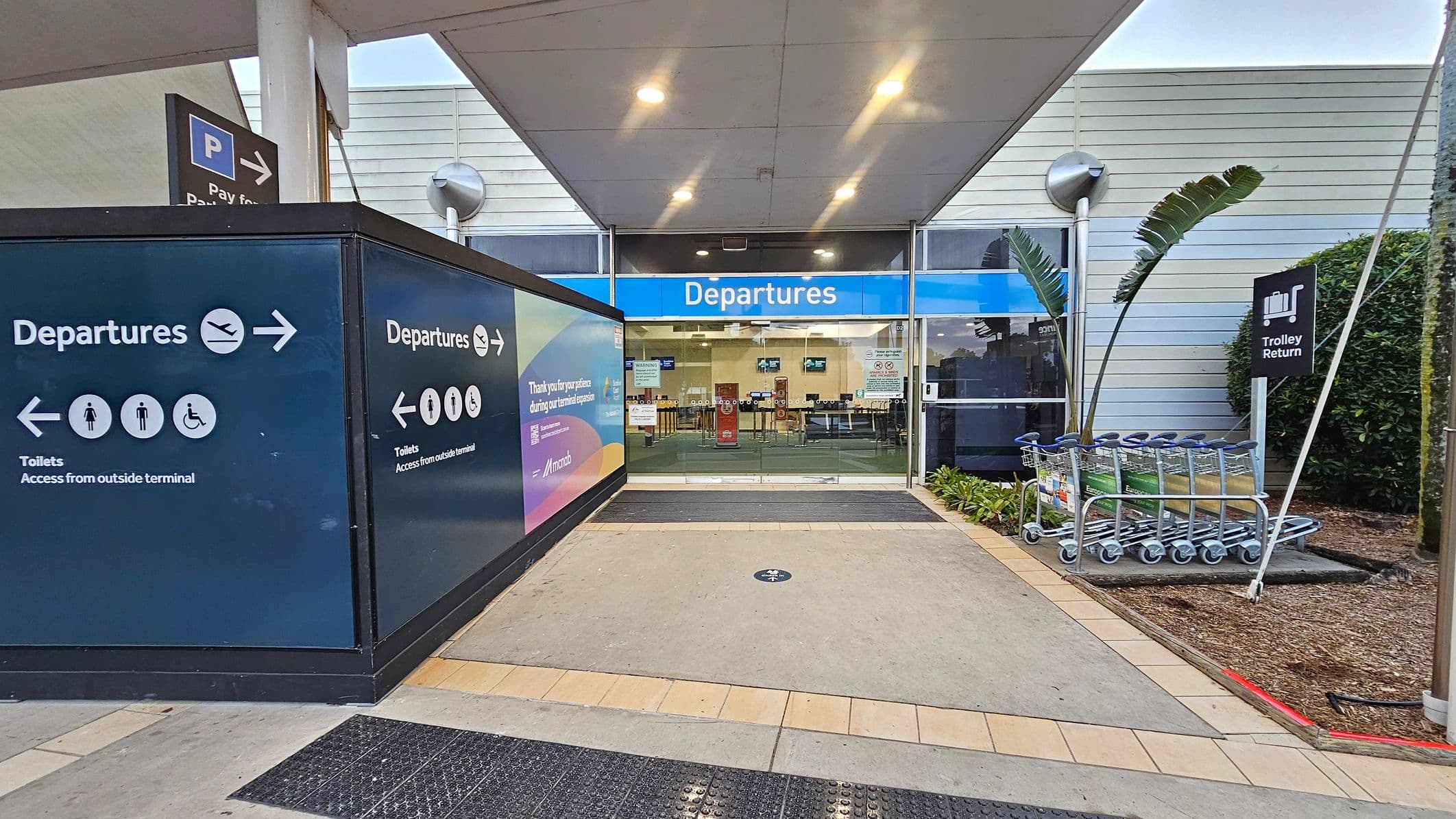 Entrance to an airport departures area. On the left, a dark blue sign with white text and icons directs to departures and toilets. In the centre, glass doors with a blue "Departures" sign above lead inside. On the right, a row of luggage trolleys is parked near a "Trolley Return" sign. The floor is a mix of tiles and concrete, and the ceiling is white with recessed lighting. A plant with large leaves is near the trolleys.