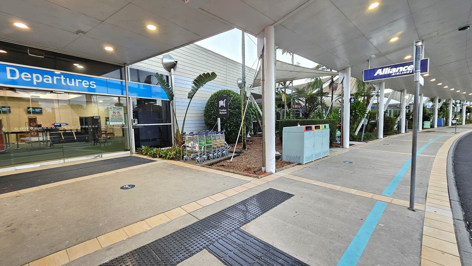 Entrance to an airport terminal with a blue "Departures" sign above glass sliding doors on the left. A row of luggage trolleys is parked near a "Trolley Return" sign. The right side features a covered walkway with a sign for "Alliance Airlines" and a row of rubbish bins. The ground is paved with a mix of concrete and tiles, and there are plants and small trees along the building's exterior. The ceiling is lined with lights.