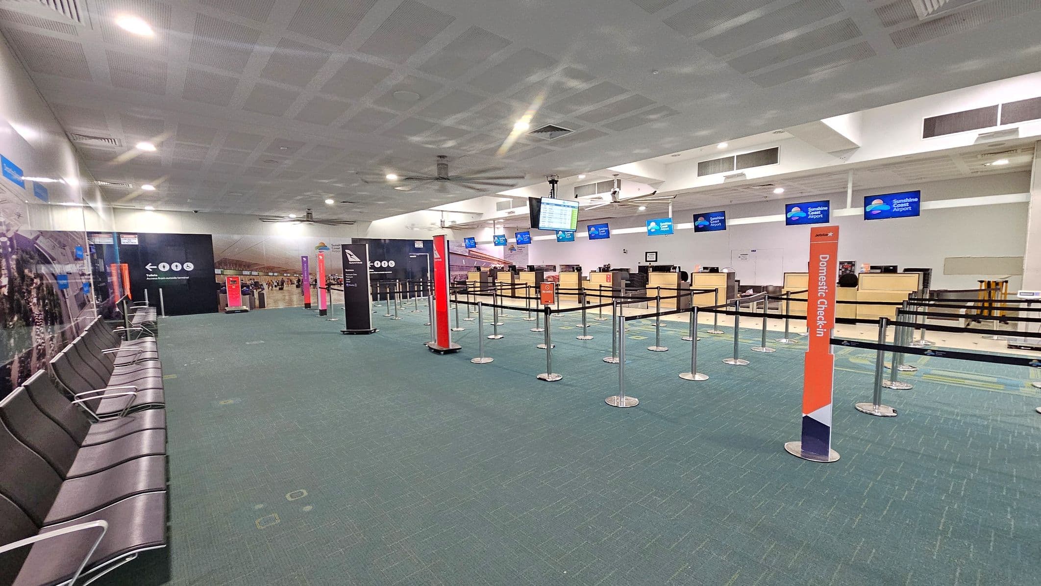 An airport check-in area with a green carpeted floor. On the right, there are several check-in counters with digital screens above them displaying "Sunshine Coast Airport". In the centre, there are queue barriers with signs indicating "Domestic Check-in". On the left, there is a row of black seating against a wall with informational displays. The ceiling is white with recessed lighting and ventilation panels.