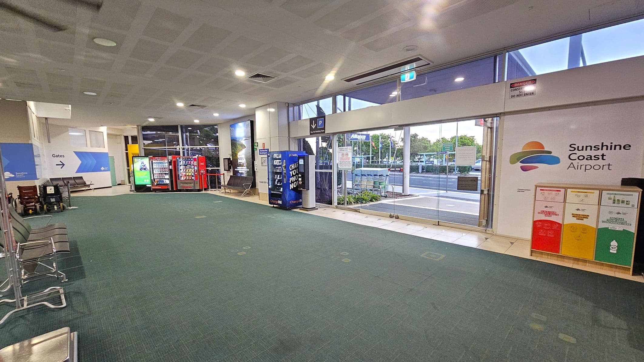 Inside an airport terminal with a green and grey carpeted floor. On the left, there are rows of metal chairs and a sign pointing to the gates. In the centre, vending machines are lined up against the wall. On the right, large glass doors lead outside, with a sign for Sunshine Coast Airport on the wall. Recycling bins in red, yellow, and green are positioned nearby. The ceiling is light-coloured with recessed lighting.
