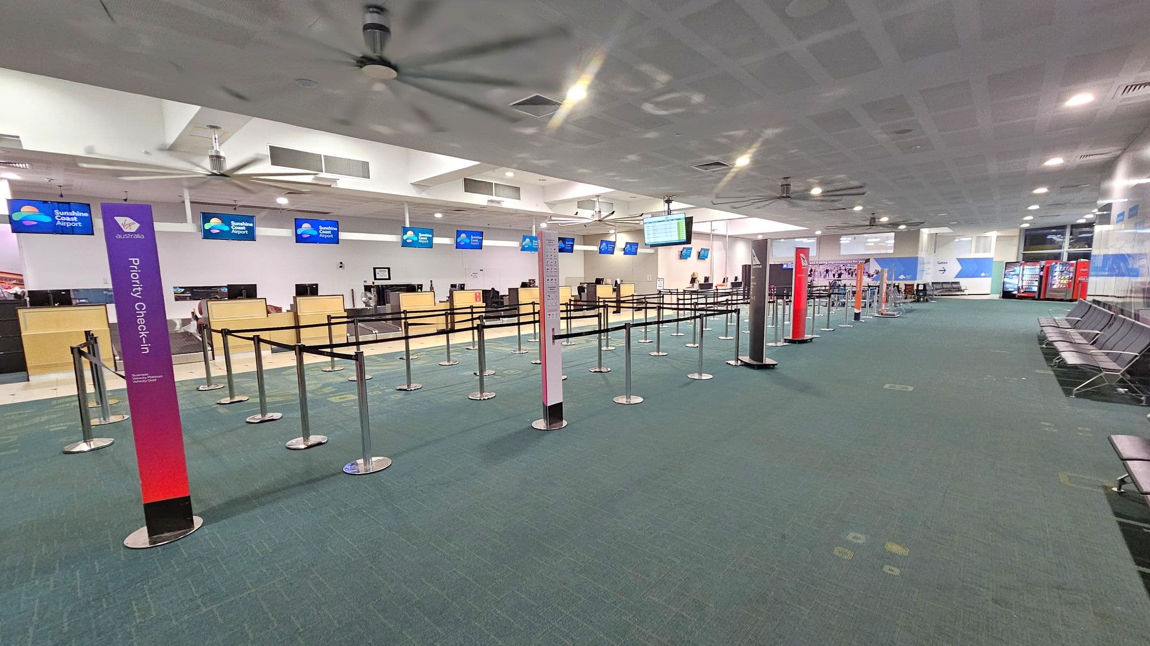 An airport check-in area with a green carpeted floor. On the left, there are check-in counters with digital screens displaying "Sunshine Coast Airport." In the centre, there are queue barriers with signs for priority check-in. On the right, there are rows of black seating and vending machines. The ceiling is white with large fans and recessed lighting.