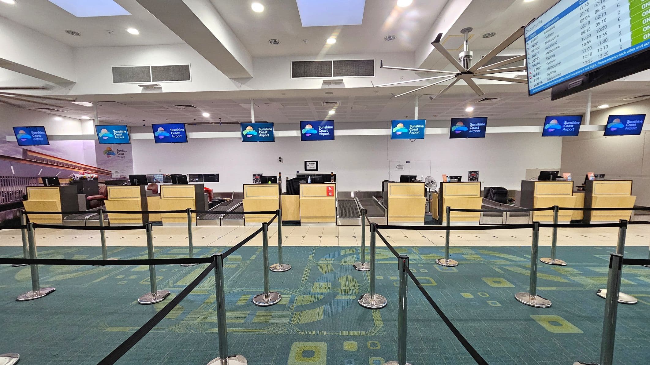 A row of check-in counters at an airport, each with a monitor displaying "Sunshine Coast Airport". The counters are wooden with black tops, and there are black stanchions with belts forming a queue area in front. The floor is carpeted with a green and blue geometric pattern. Above, a large ceiling fan is visible, and a digital flight information screen is on the right. The ceiling is white with recessed lighting and skylights.