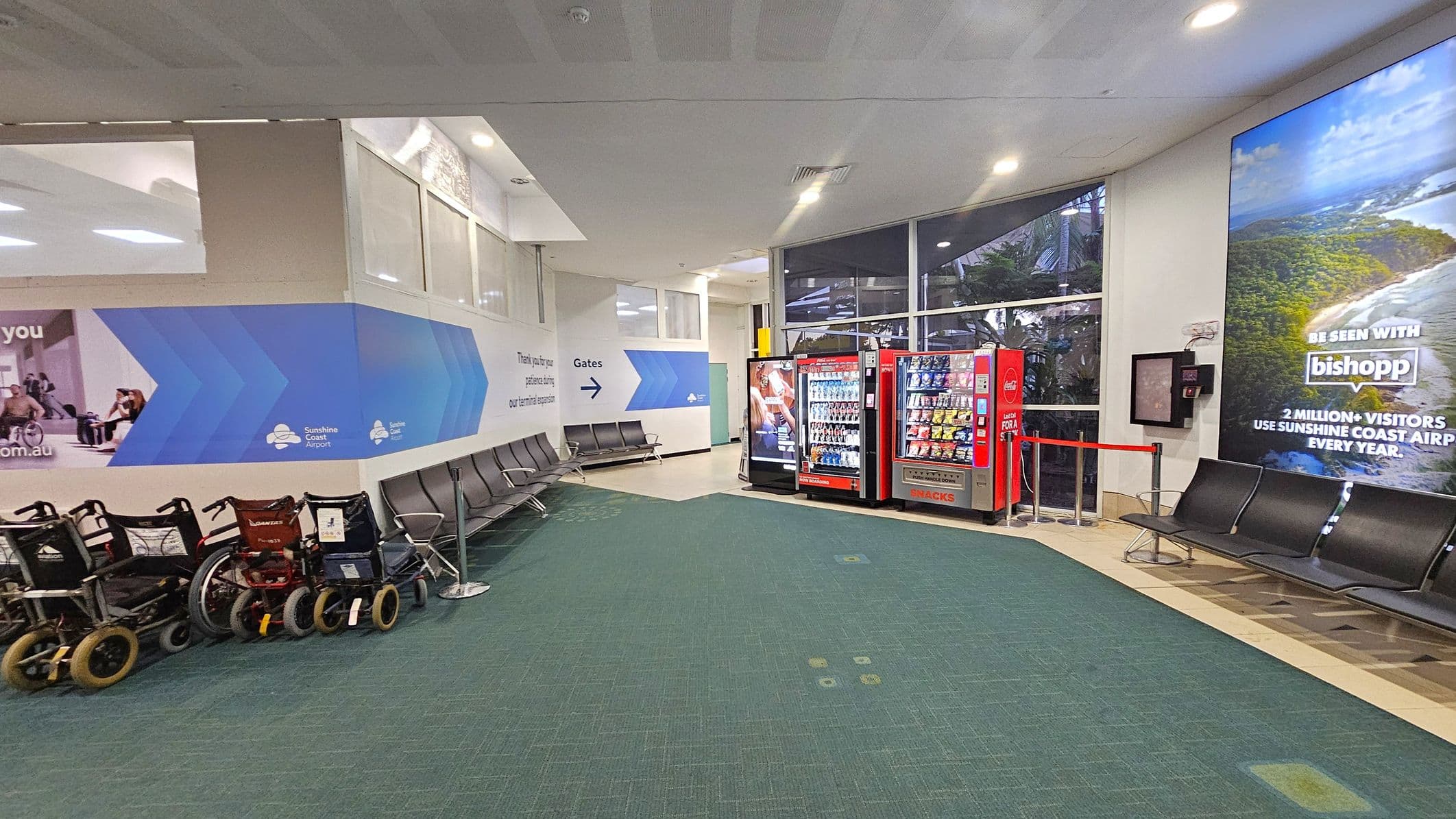 On the left, there are several wheelchairs lined up against a wall with a blue and white design featuring the Sunshine Coast Airport logo. Adjacent to them are rows of black seating. In the centre, the floor is covered with green carpet leading to a corner with vending machines. On the right, more seating is visible beneath a large advertisement showing a coastal scene. The ceiling is white with recessed lighting, and large windows in the background allow natural light to enter.