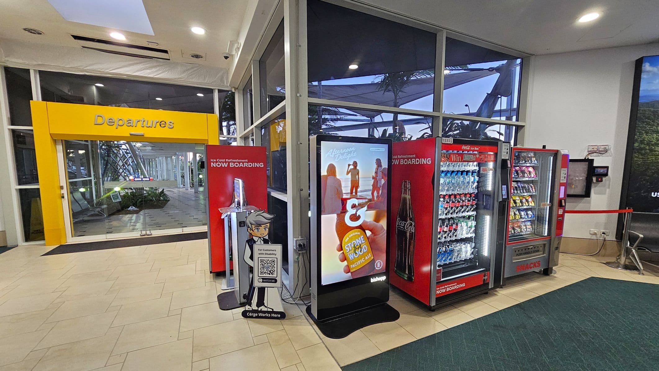 A brightly lit airport area with a yellow "Departures" sign above glass doors on the left. In the centre, there's a digital display with an advertisement, flanked by two red vending machines stocked with drinks and snacks. A stand with a cartoon character and a QR code is in front of the display. The floor is tiled in a light colour, and the ceiling has recessed lighting. Large windows in the background reveal some outdoor structures and plants.